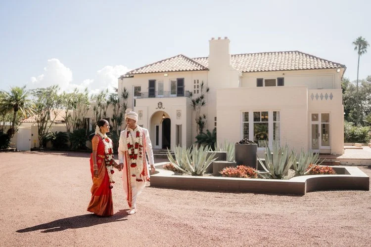 A couple dressed in traditional Indian wedding attire walking in front of a large white house with desert plants in the foreground.