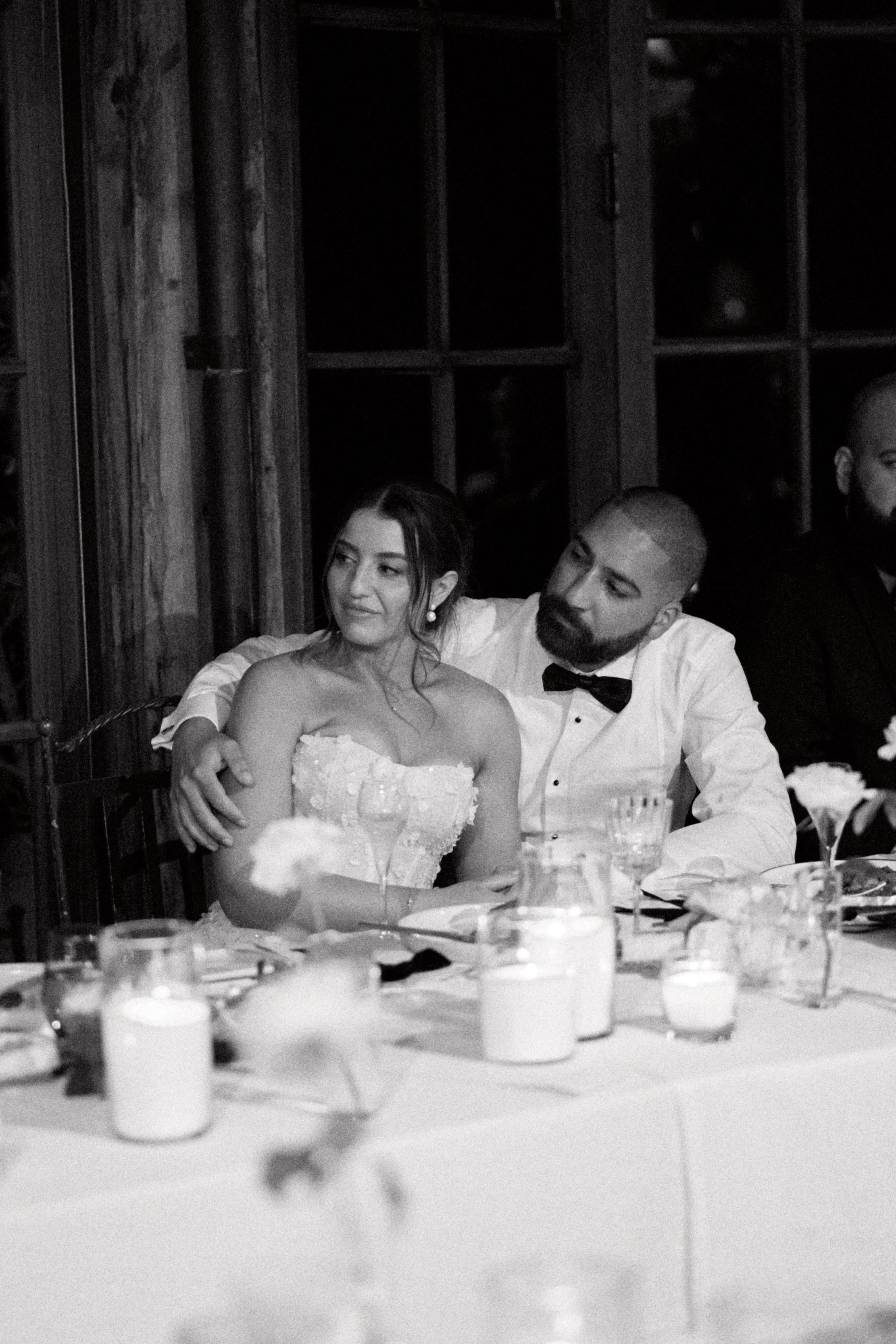A black and white photo of a bride and groom sitting together at a wedding reception table. The bride is wearing a strapless dress and the groom is in a tuxedo with a bowtie. They are seated in front of a window with wooden framing, surrounded by dec
