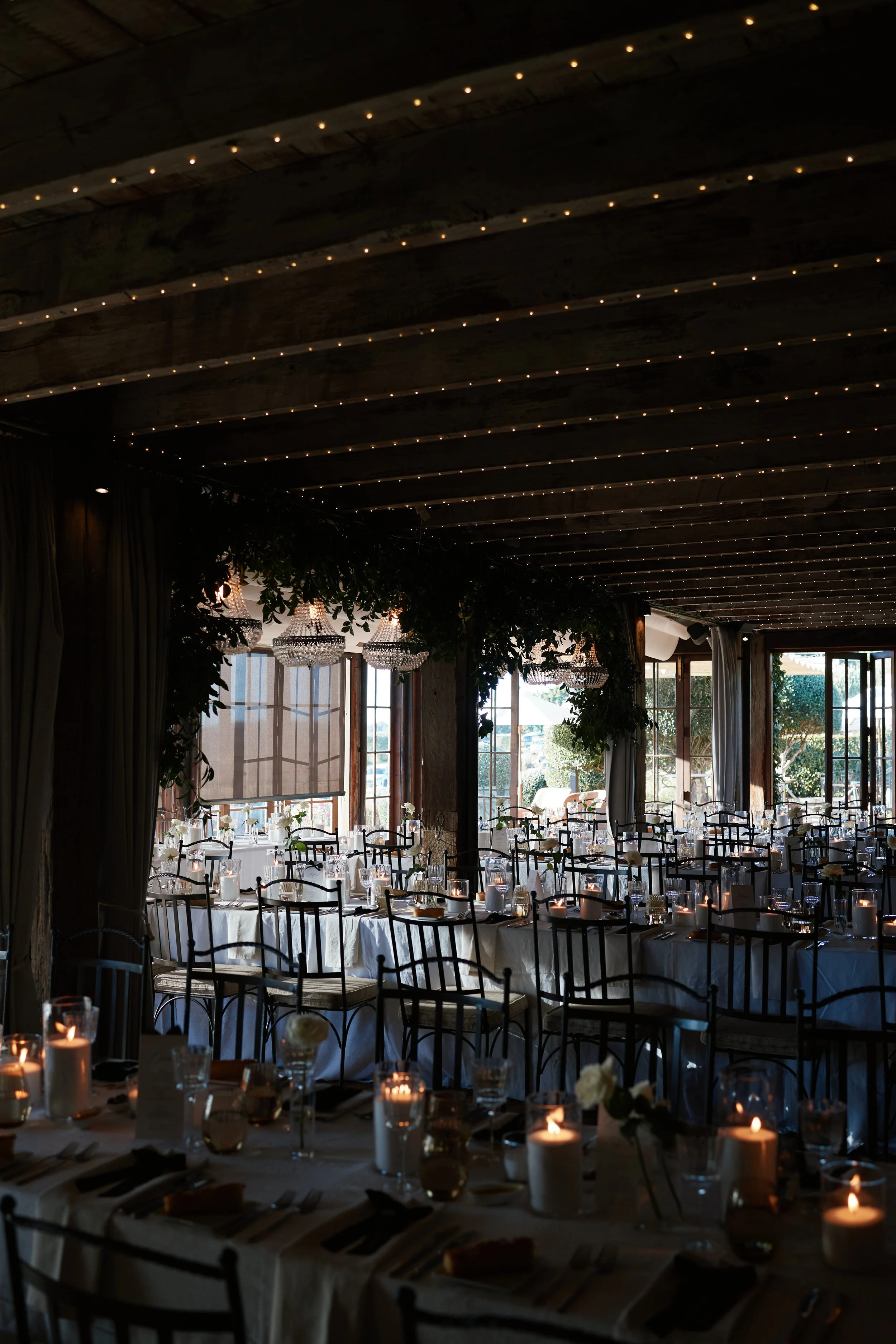 Elegant reception hall with long dining tables set for an event, decorated with candles, flowers, and tableware, with large windows allowing sunlight to illuminate the space, chandeliers hanging from the ceiling, and string lights for ambiance.
