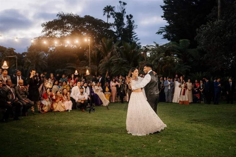 A bride and groom dancing at their outdoor wedding reception with guests seated and standing around them, trees in the background, and string lights overhead during dusk.