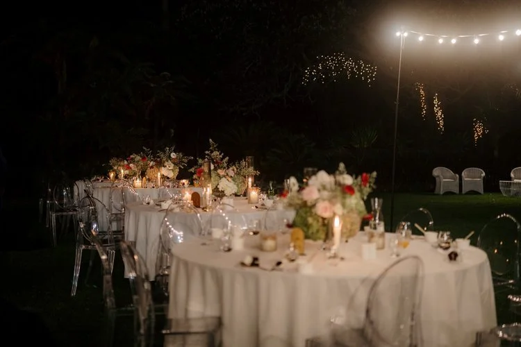 Evening outdoor wedding reception with decorated tables, floral centerpieces, candles, string lights, and white chairs under a dark sky.
