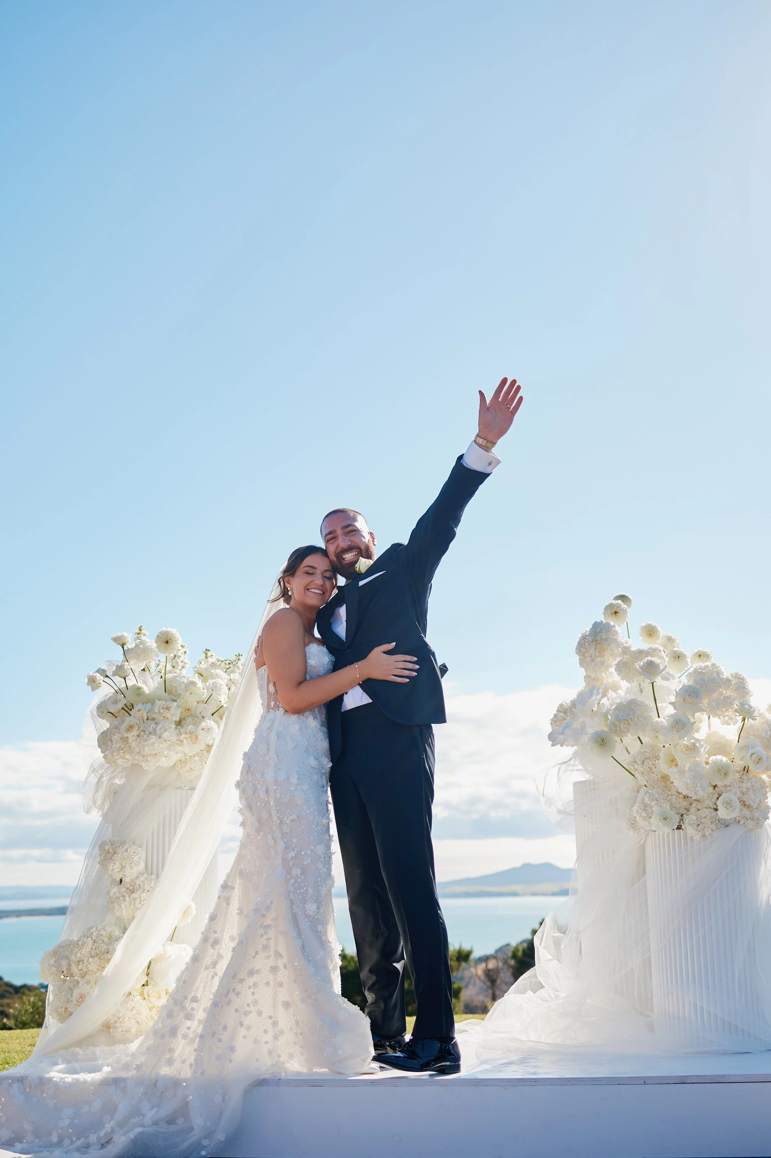 A bride and groom celebrating on their wedding day outdoors with a view of water and mountains in the background, surrounded by white floral arrangements.