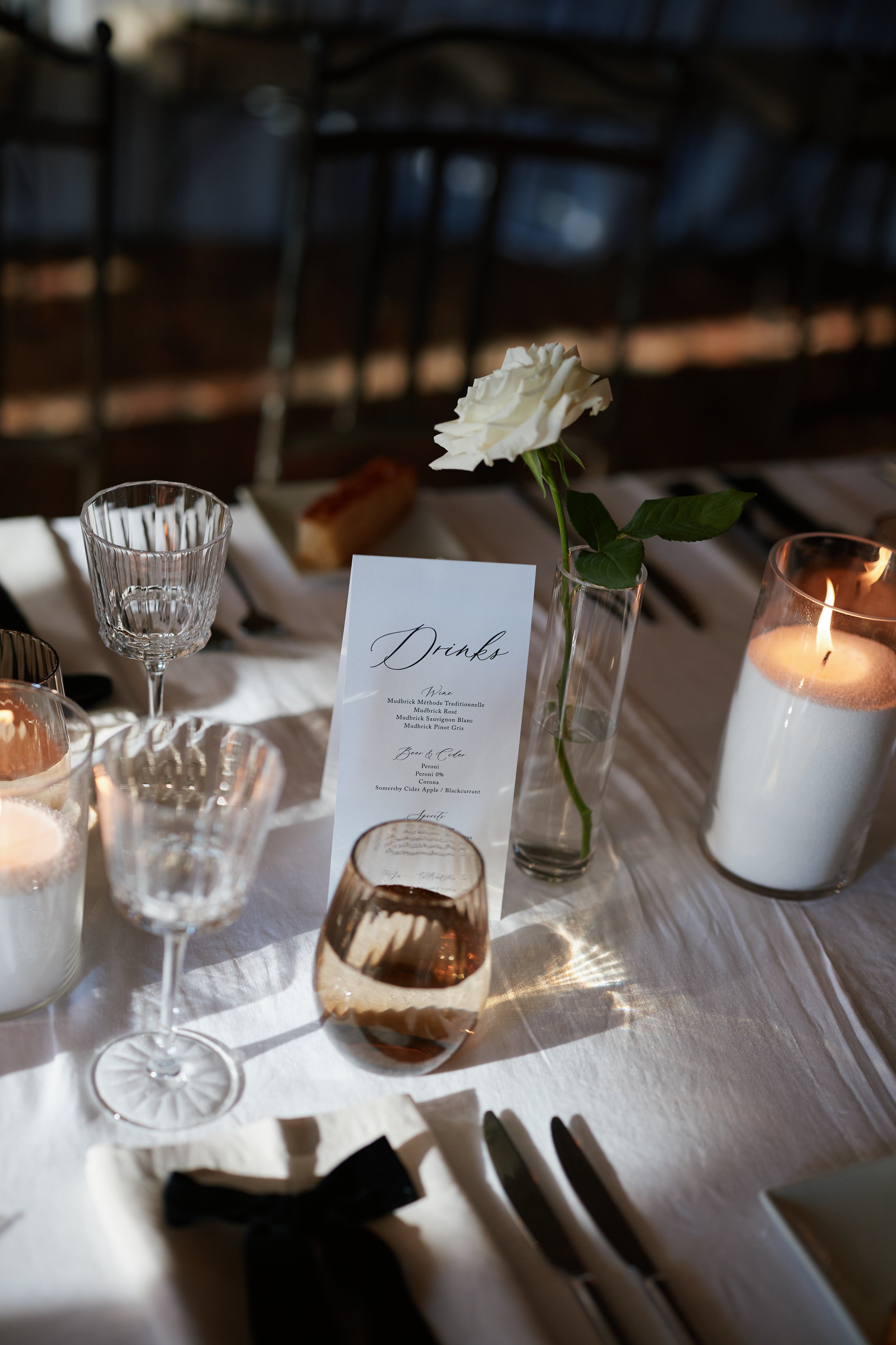 Elegant dinner table setting with a white rose in a glass vase, candles, wine glasses, a menu card, and silverware, illuminated by warm candlelight.