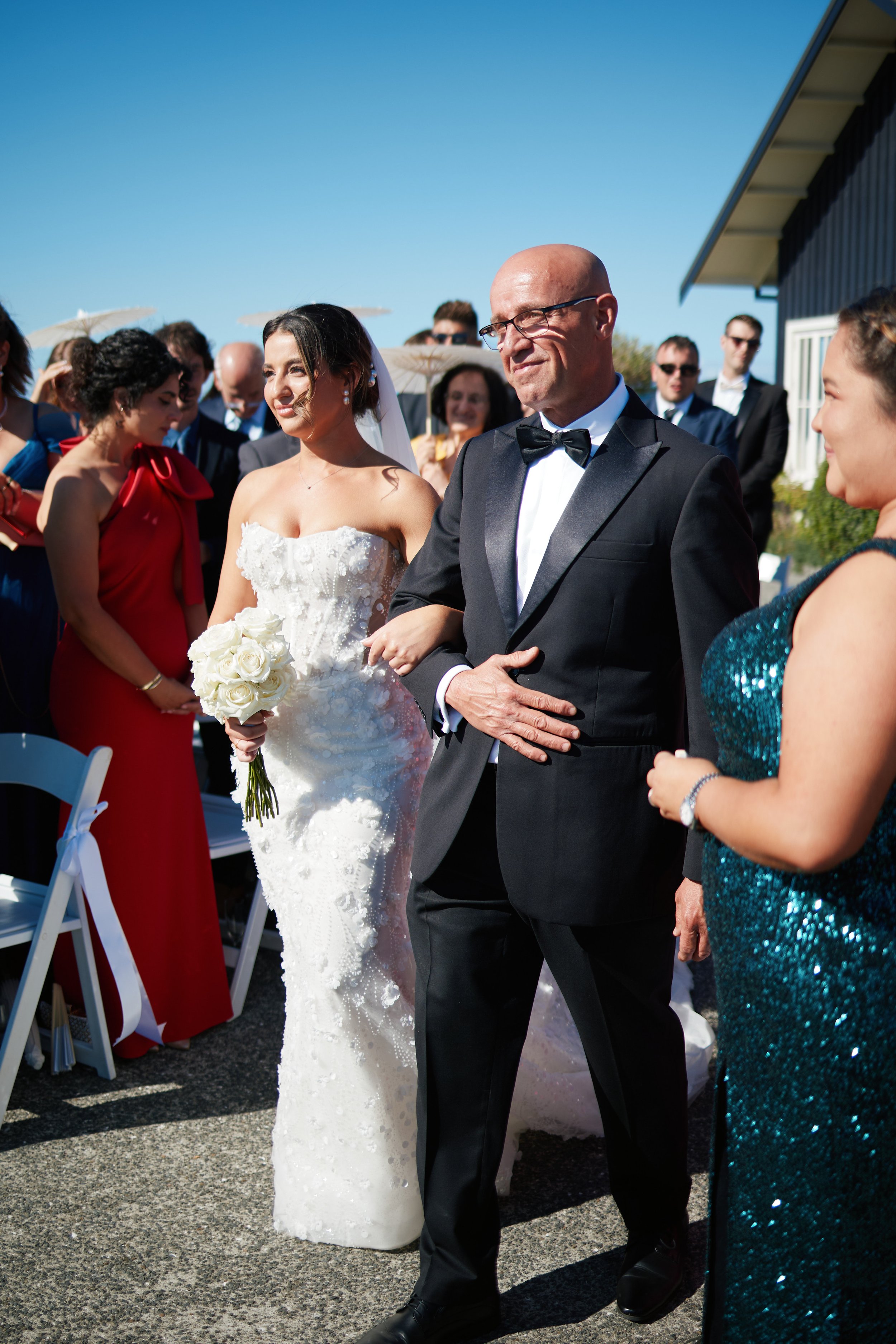 A bride in a white wedding gown holding a bouquet of white roses walking down the aisle with her father at an outdoor wedding ceremony. Guests in formal attire are watching on a sunny day.