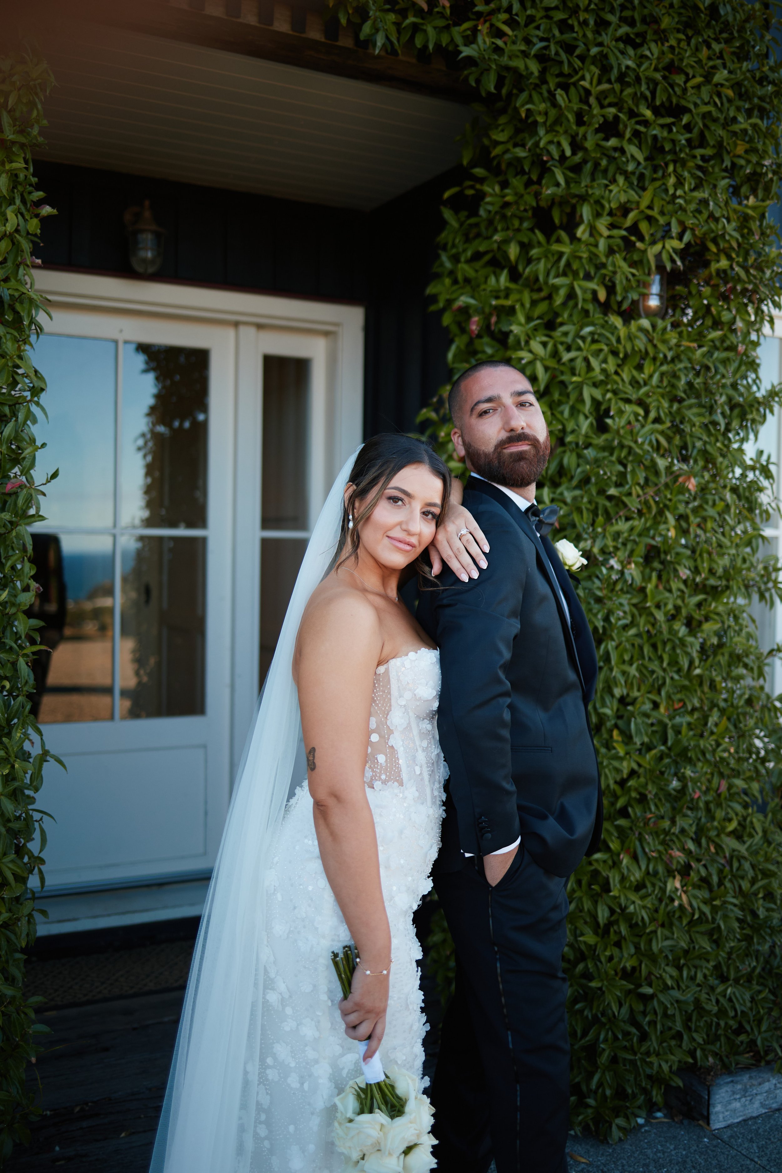 A bride and groom stand close together outside, framed by green leafy plants. The bride is holding a bouquet of white flowers and leans her head slightly on the groom's shoulder. She is dressed in a strapless white wedding gown with a long veil, and 