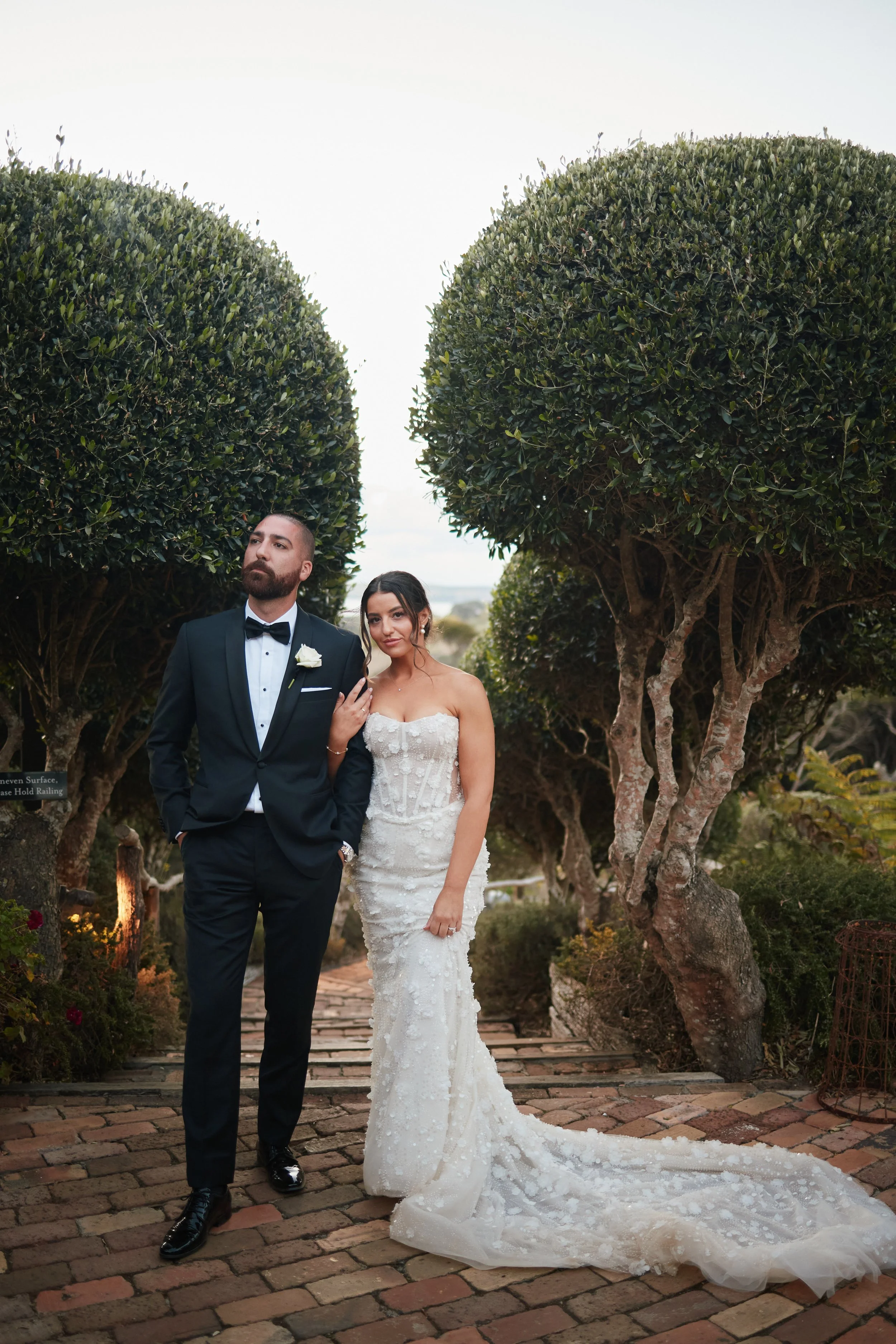 A bride and groom walking together outdoors on a brick pathway, surrounded by lush trees and greenery, dressed in wedding attire.