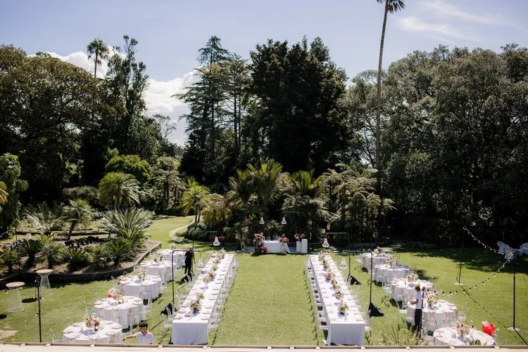 Outdoor garden wedding reception setup with long dining tables, chairs, floral centerpieces, and a table at the end, surrounded by lush greenery and tall trees.