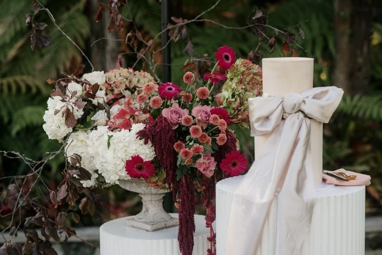 A floral arrangement featuring white hydrangeas, pink roses, and dark red blooms alongside a tall white candle with a large bow, displayed on white pedestals in a garden setting.