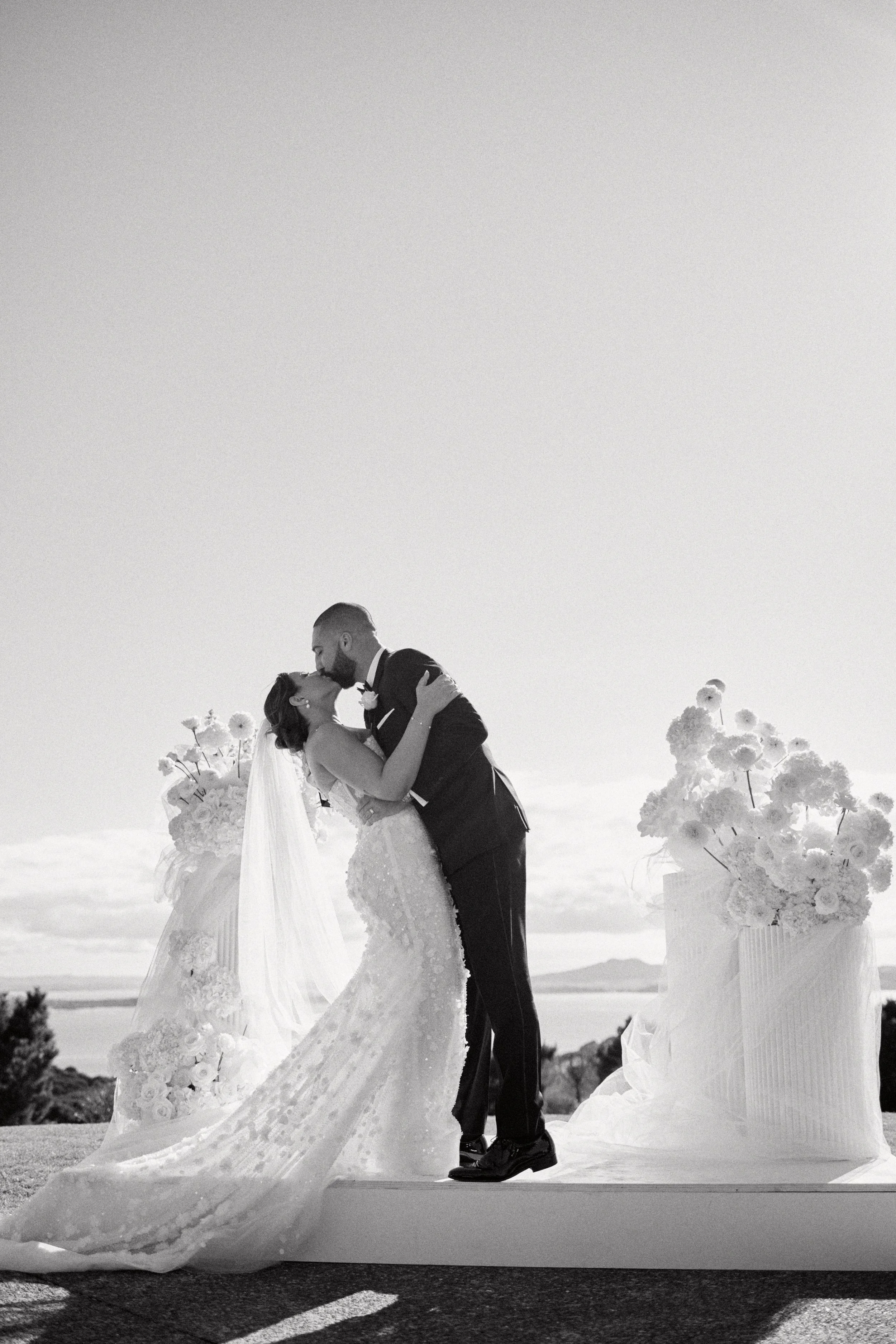 A black and white photo of a bride and groom sharing a kiss during their outdoor wedding ceremony, with large floral arrangements on pedestals and the ocean in the background.