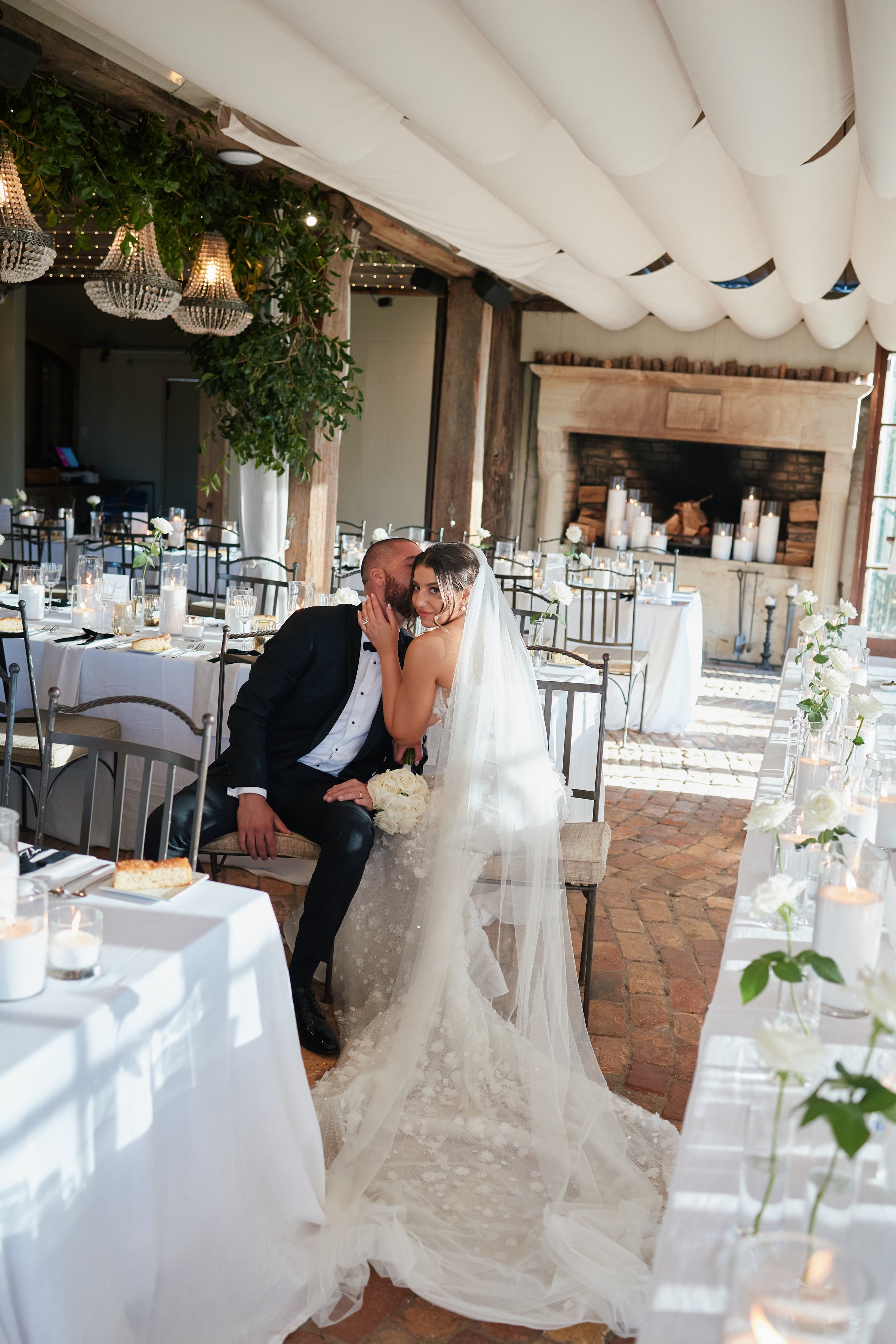 Bride and groom sitting closely at wedding reception, with the bride whispering into the groom's ear in a decorated indoor venue with tables, candles, and chandeliers.