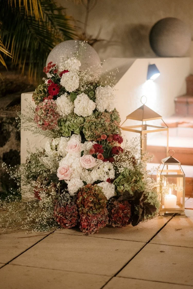 A floral arrangement with white, pink, red, and green flowers, illuminated by candle lanterns, set on a wooden surface with a decorative background.