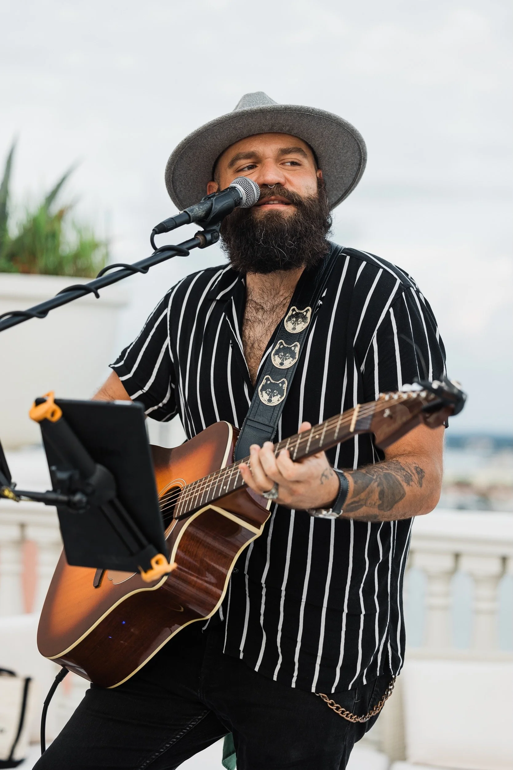 A bearded man wearing a gray wide-brimmed hat and a black and white striped shirt is playing an acoustic guitar and singing into a microphone outdoors during daytime. There are some plants and a cloudy sky in the background. Vinnie Hines