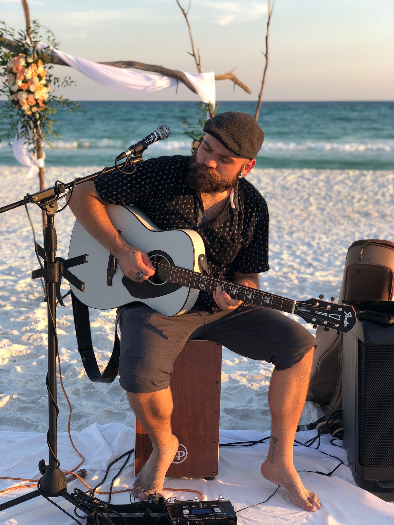 A man (Vinnie Hines) playing an acoustic guitar on a beach with sand and ocean in the background, decorated with a floral arch and white drapery, during sunset or sunrise, somewhere in Florida.
