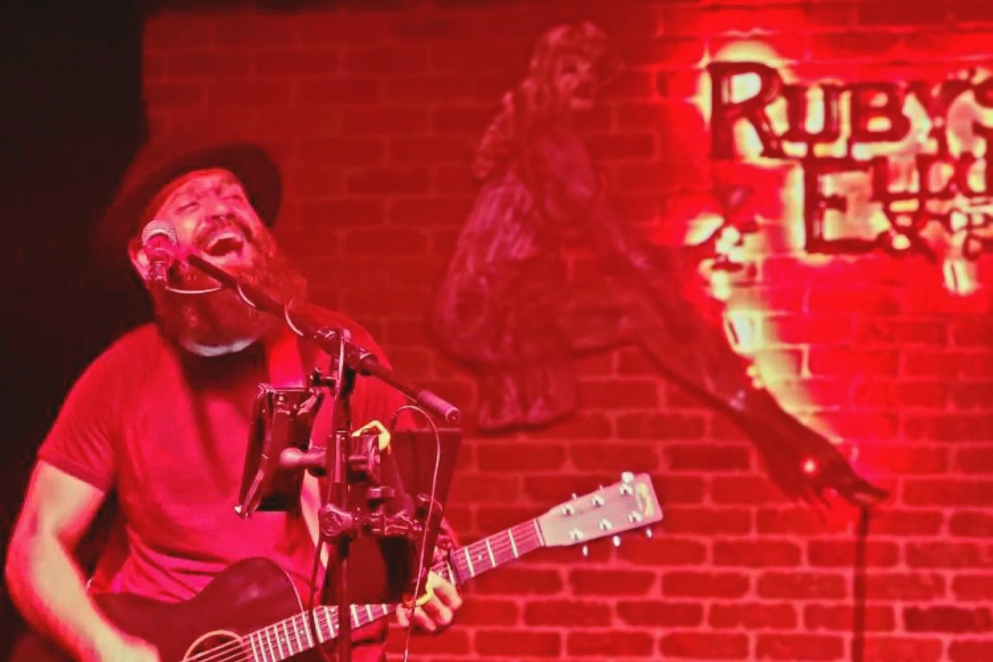 Vinnie Hines, A singer with a beard and hat singing into a microphone, playing a guitar, with a red brick wall and a neon sign in the background at Ruby's Elixir in St. Petersburg, FL.