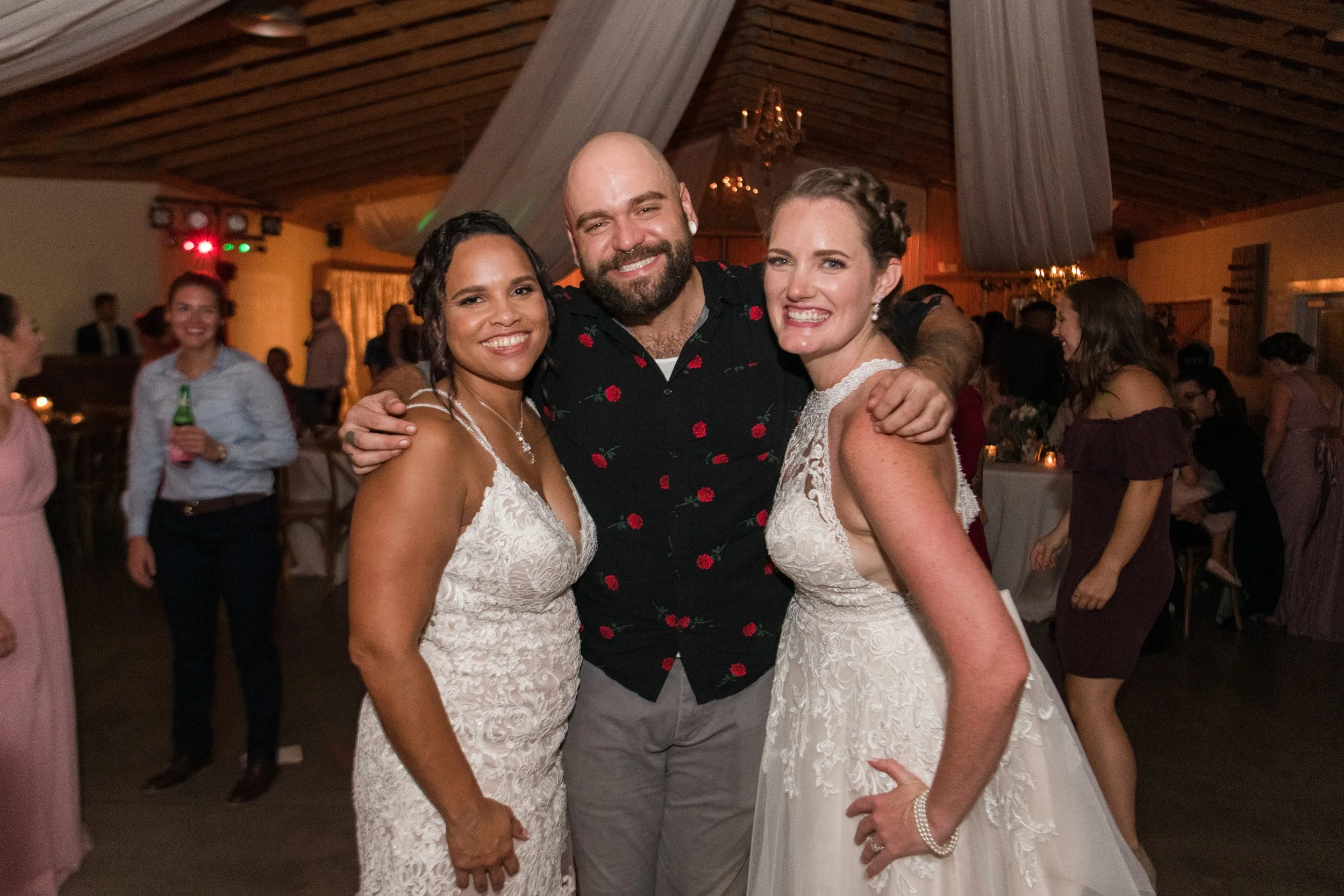 Three guests embrace at a wedding reception in a wood-paneled venue.