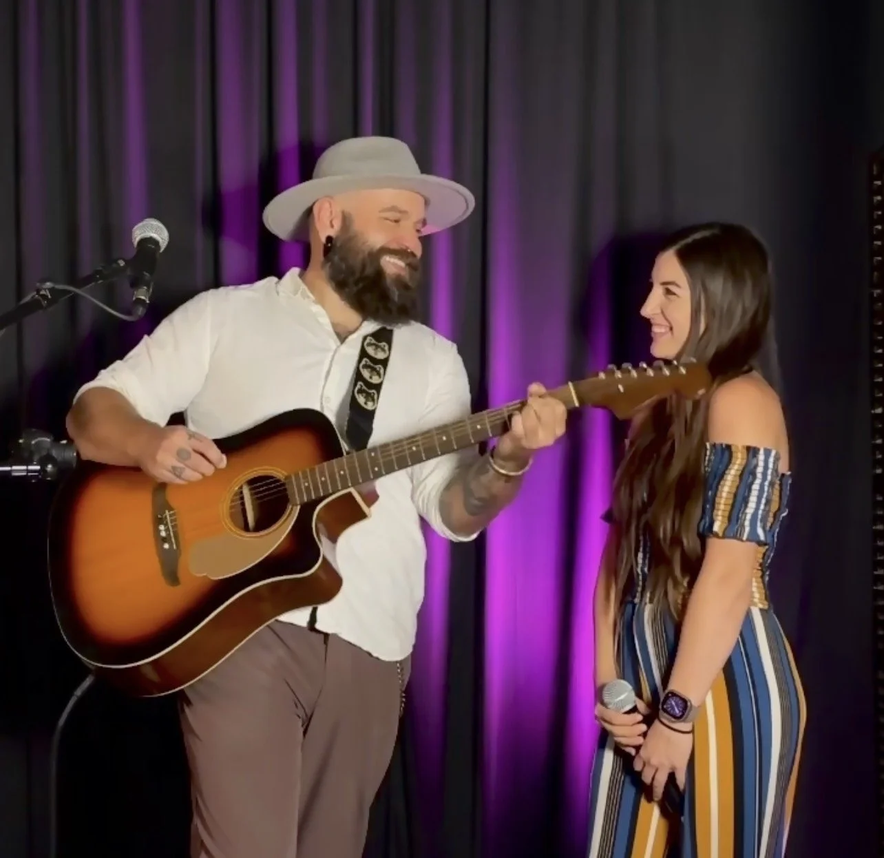 A man with a beard wearing a light gray hat and a white shirt, playing an acoustic guitar on stage, smiling at a young woman with long brown hair wearing a colorful striped dress, who is holding a microphone and smiling back, with purple stage curtains behind them.