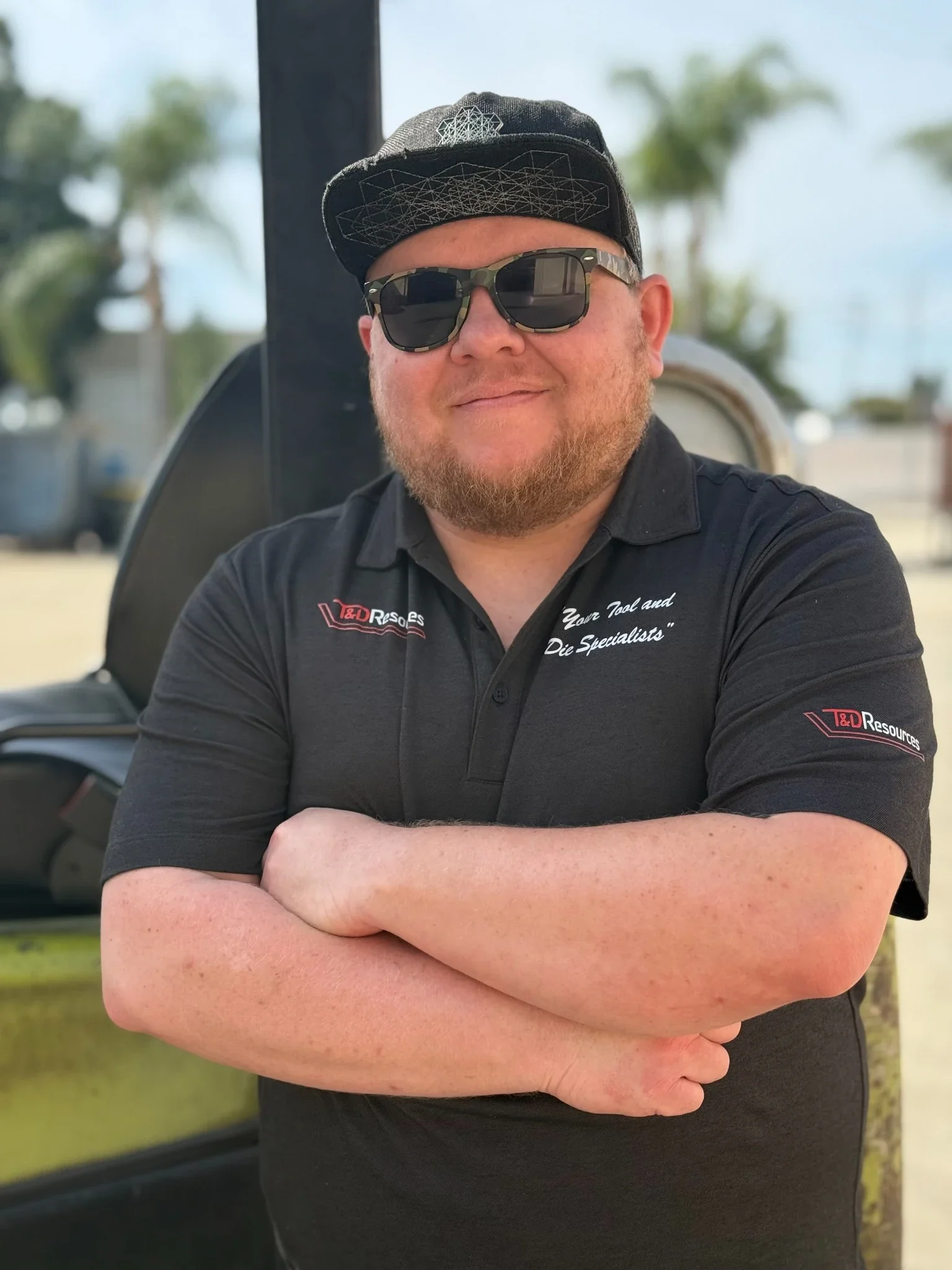 Alex from T&D Resources standing in front of shop equipment, wearing a black T&D Resources polo with arms crossed