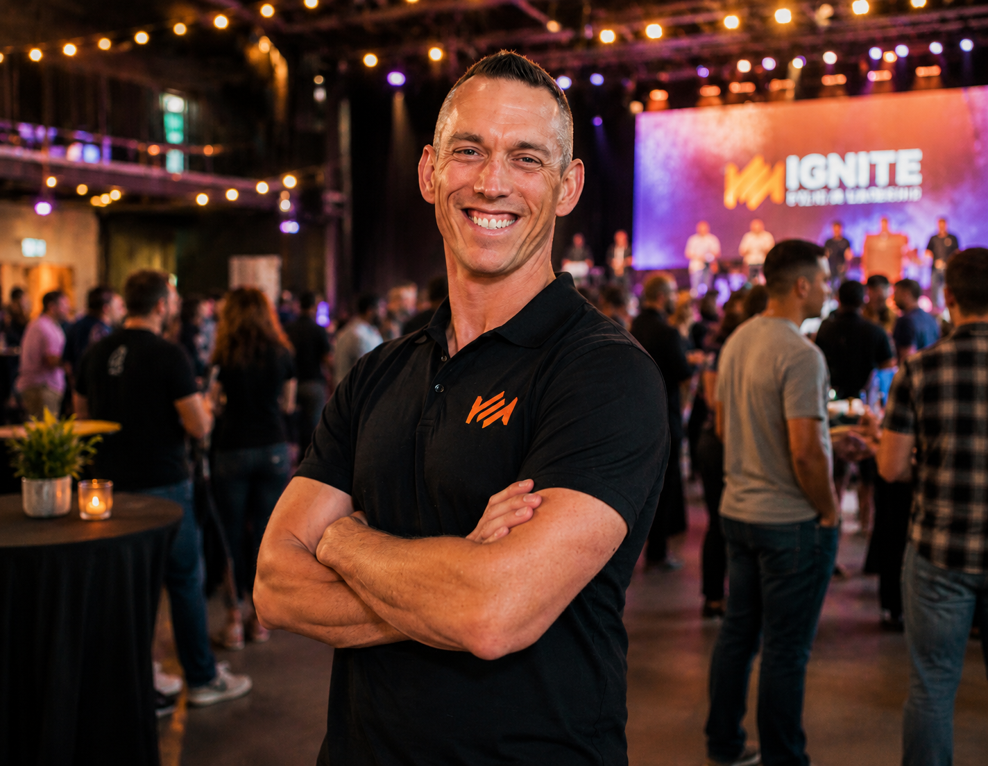 A smiling man with short hair, wearing a black shirt with an orange logo, standing with arms crossed in a crowded event space with a stage in the background displaying the word 'IGNITE' and various people mingling.
