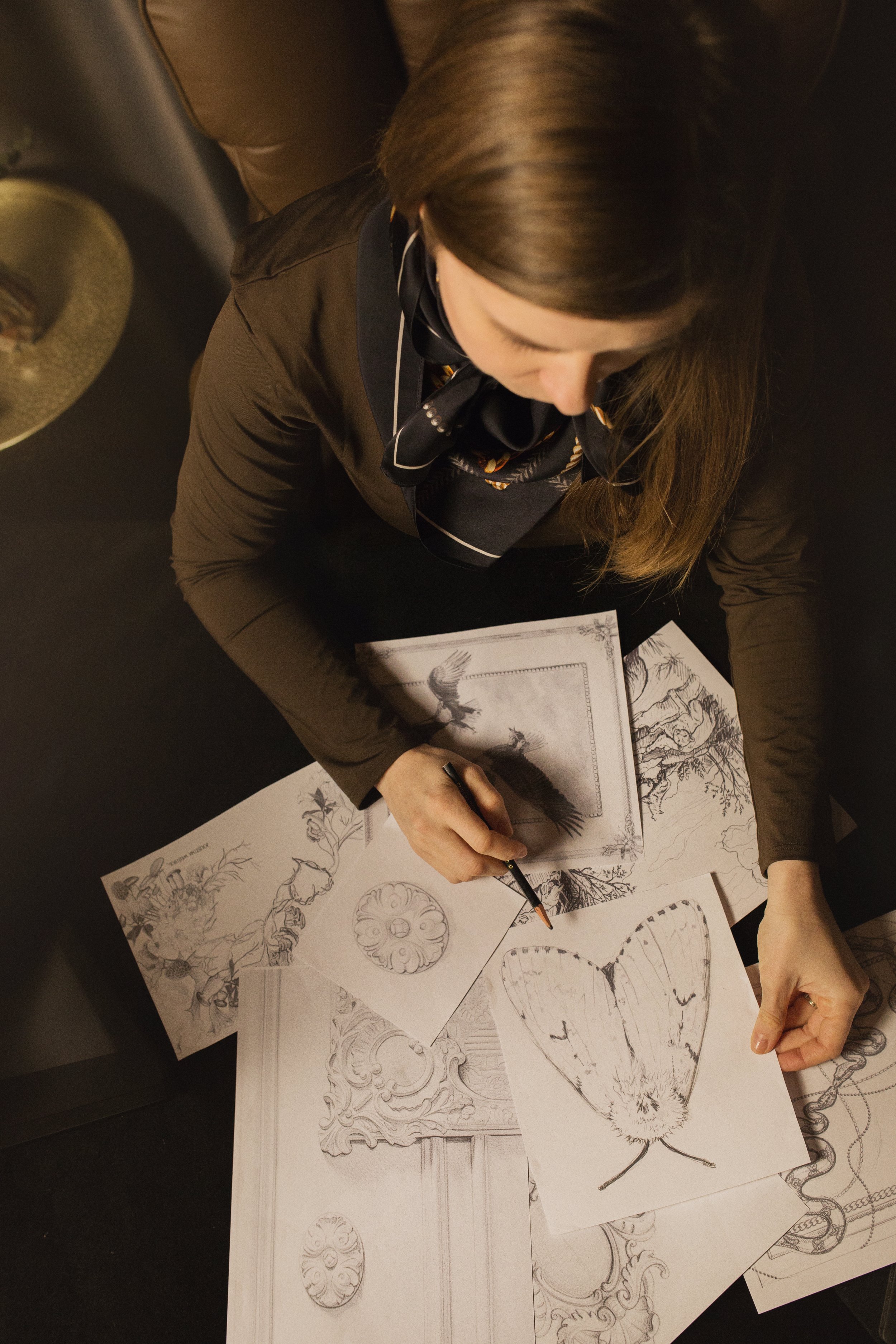 A woman sitting at a table drawing and sketching various artistic designs and illustrations, including a butterfly, a bird, and decorative patterns.