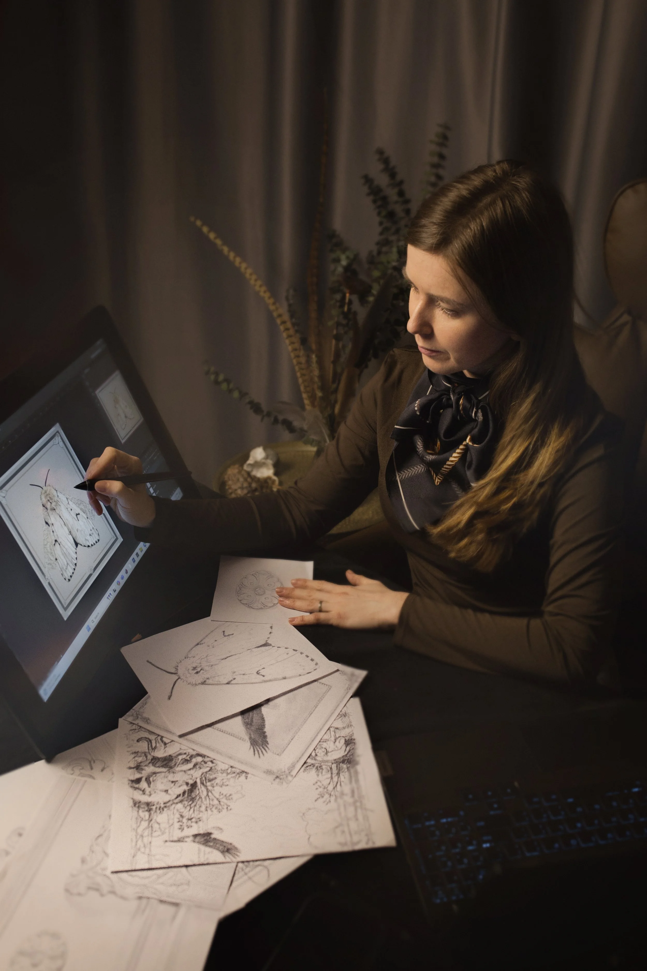 A woman working on butterfly and bird illustrations using a digital drawing tablet, surrounded by printed sketches and drawings of nature scenes.