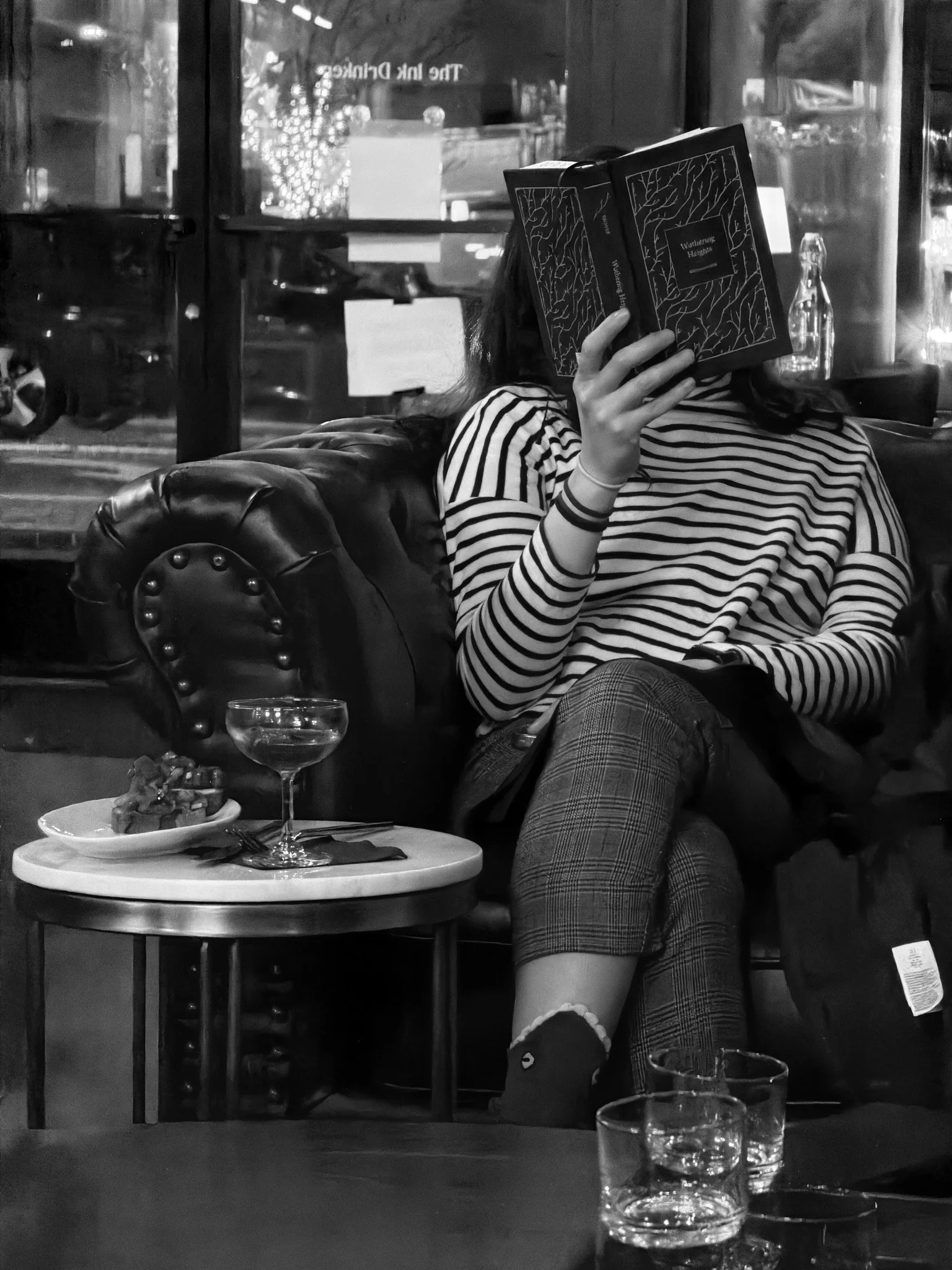 Black and white photo of a woman in a bar, sitting on a leather couch reading a book.