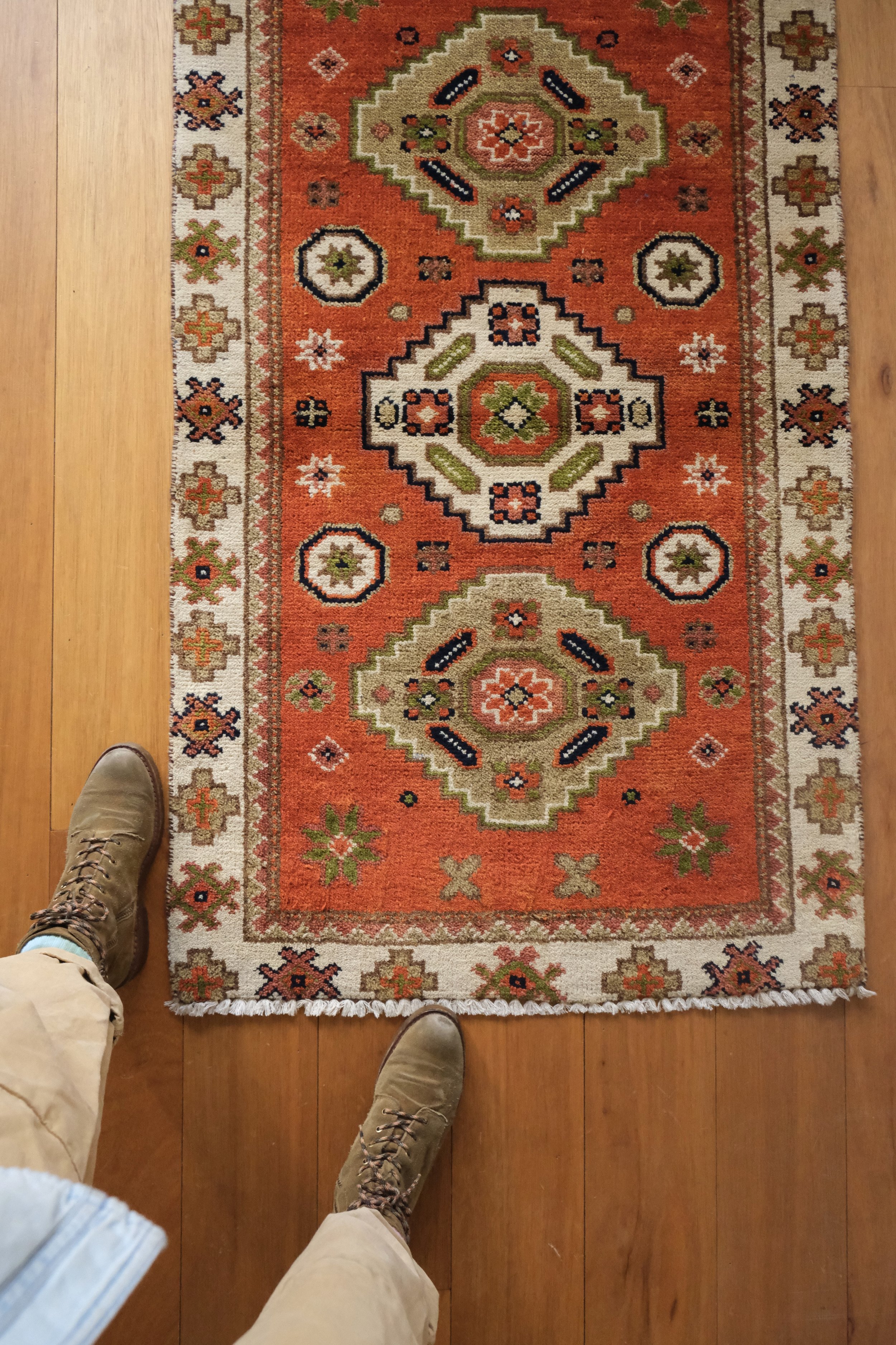 a closer up overhead photo of the vintage Turkish tribal rug with Annas feet for scale, a thin border with repeating pattern and a lovely persimmon field with geometric triple medallion design