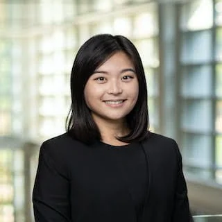 Veronica Kang smiles, wearing a black shirt and suit jacket in front of bright windows.