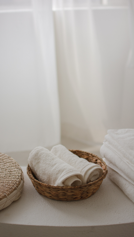 Stacked white towels and rolled white hand towels in a wicker basket on a white surface.
