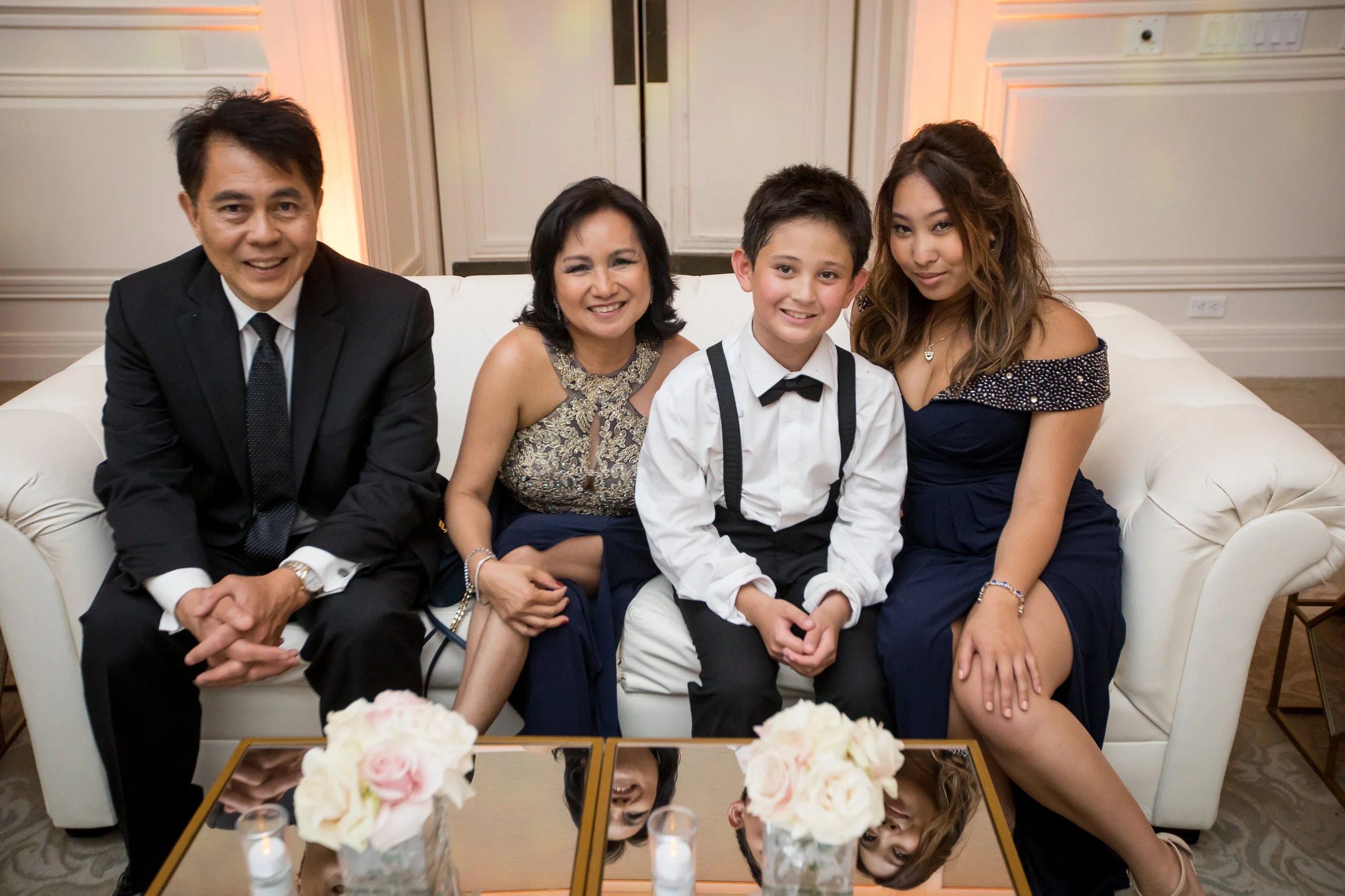 A group of five people sitting on a white sofa at a formal event, including two men in black suits, a woman in a gold and black dress, a young boy in a white shirt and suspenders, and a young woman in a blue dress, with a table of white flowers and c
