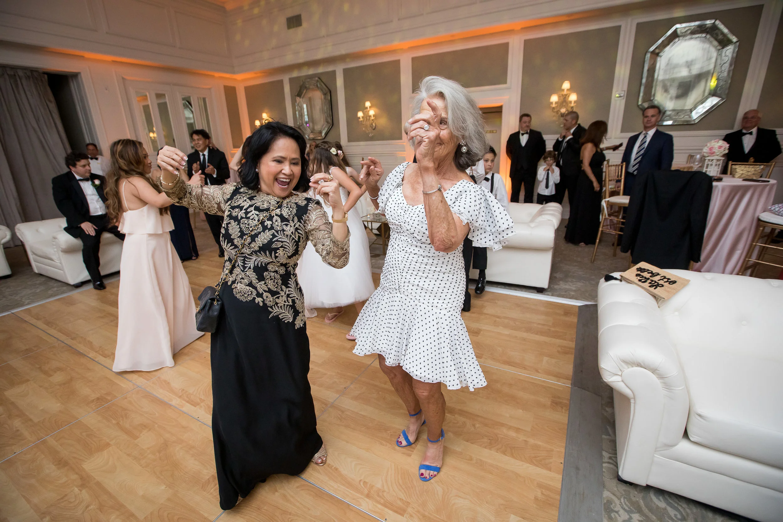 Two women dancing and having fun at a celebration or wedding reception, with guests in formal attire in the background.