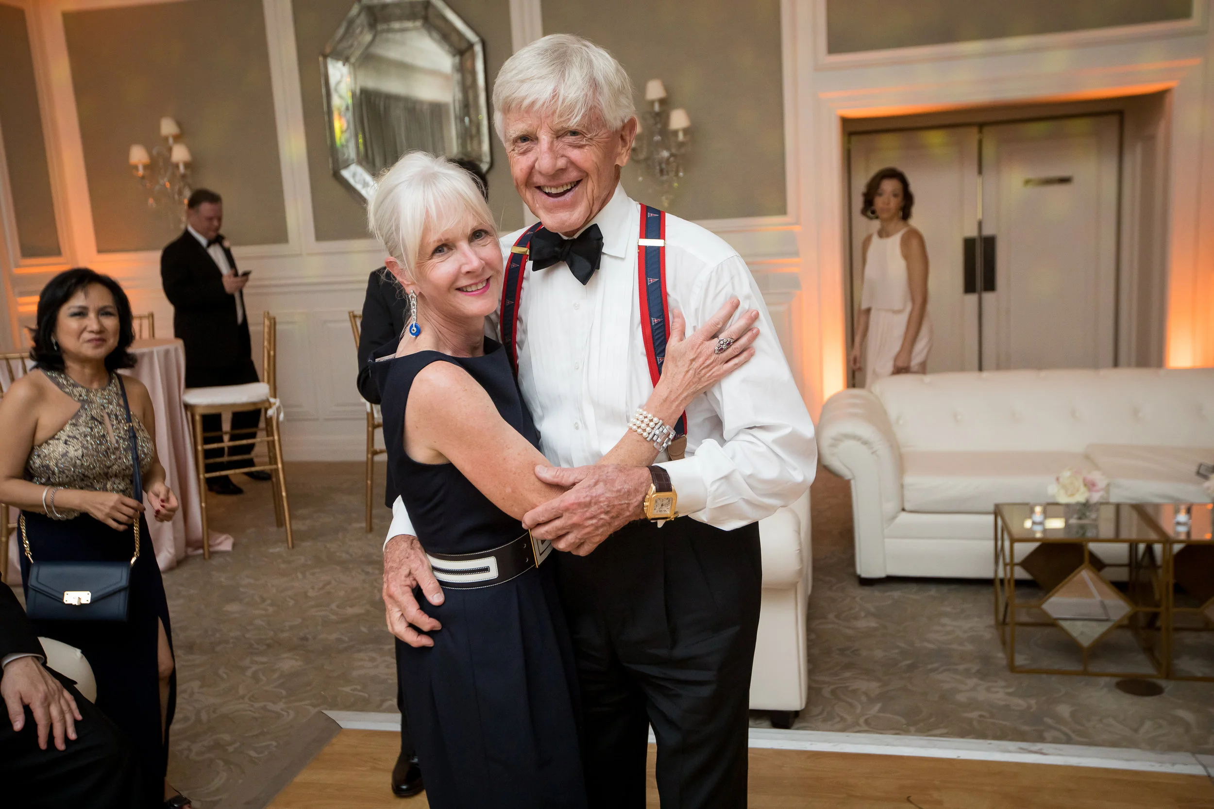 An elderly couple dancing and smiling at a formal event or party, with other guests in the background in an elegant room decorated with mirrors and warm lighting.