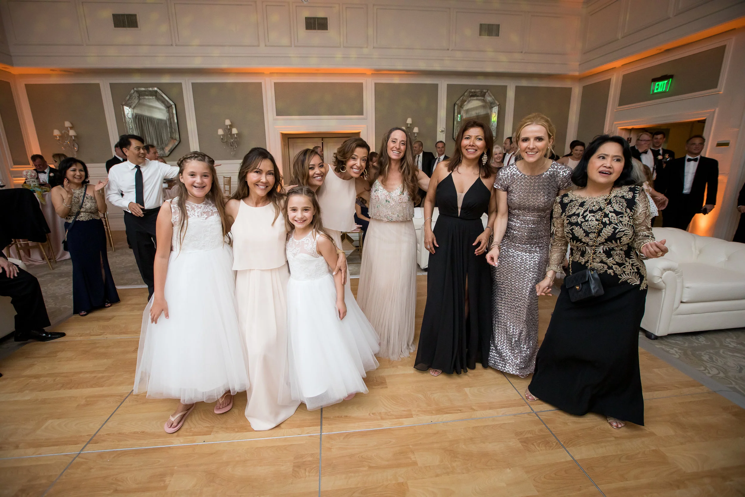 Group of women and young girls dressed in formal attire at a celebration or wedding reception, standing together and smiling.