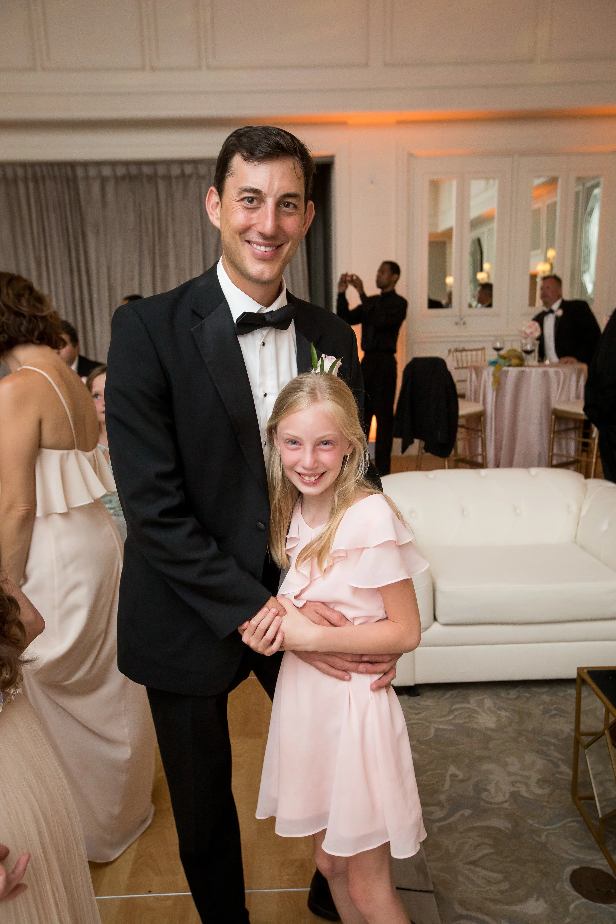 A man in a tuxedo and a girl in a pink dress smiling and holding hands at a formal event or celebration.