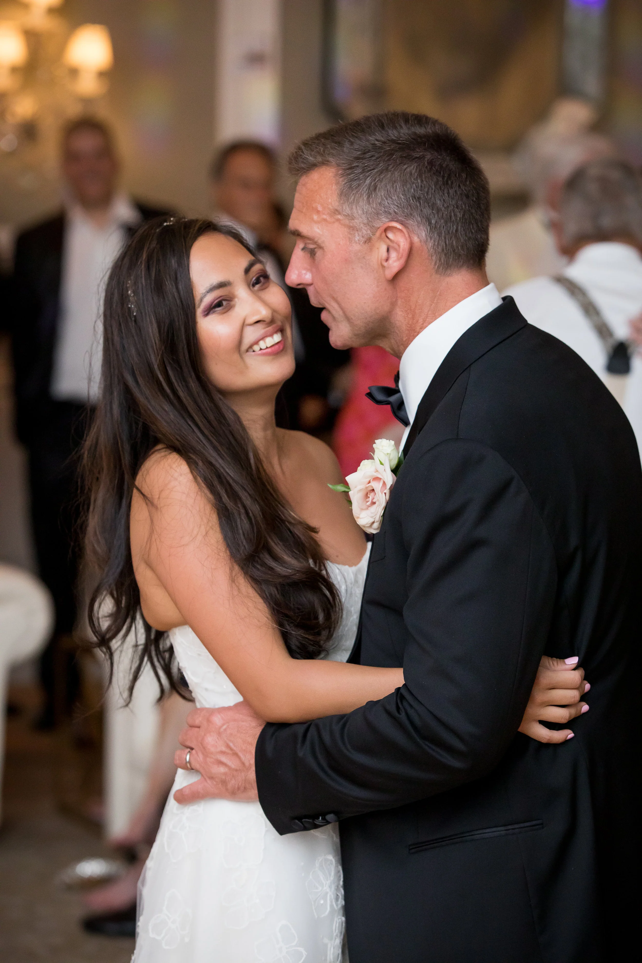 A bride and groom dance closely at their wedding reception, surrounded by guests in formal attire.