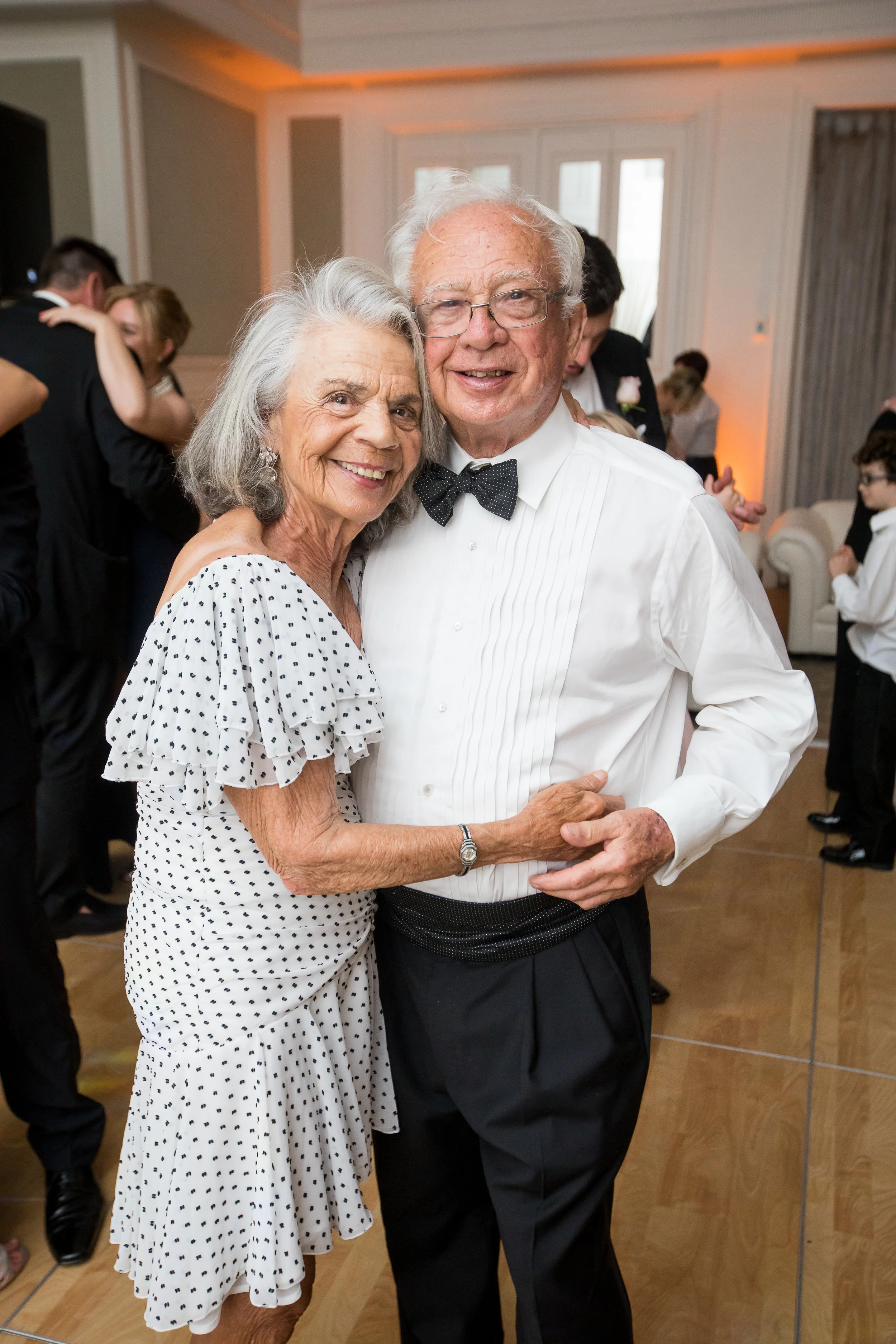 An elderly couple dancing and smiling at a formal event, surrounded by other people in elegant attire.