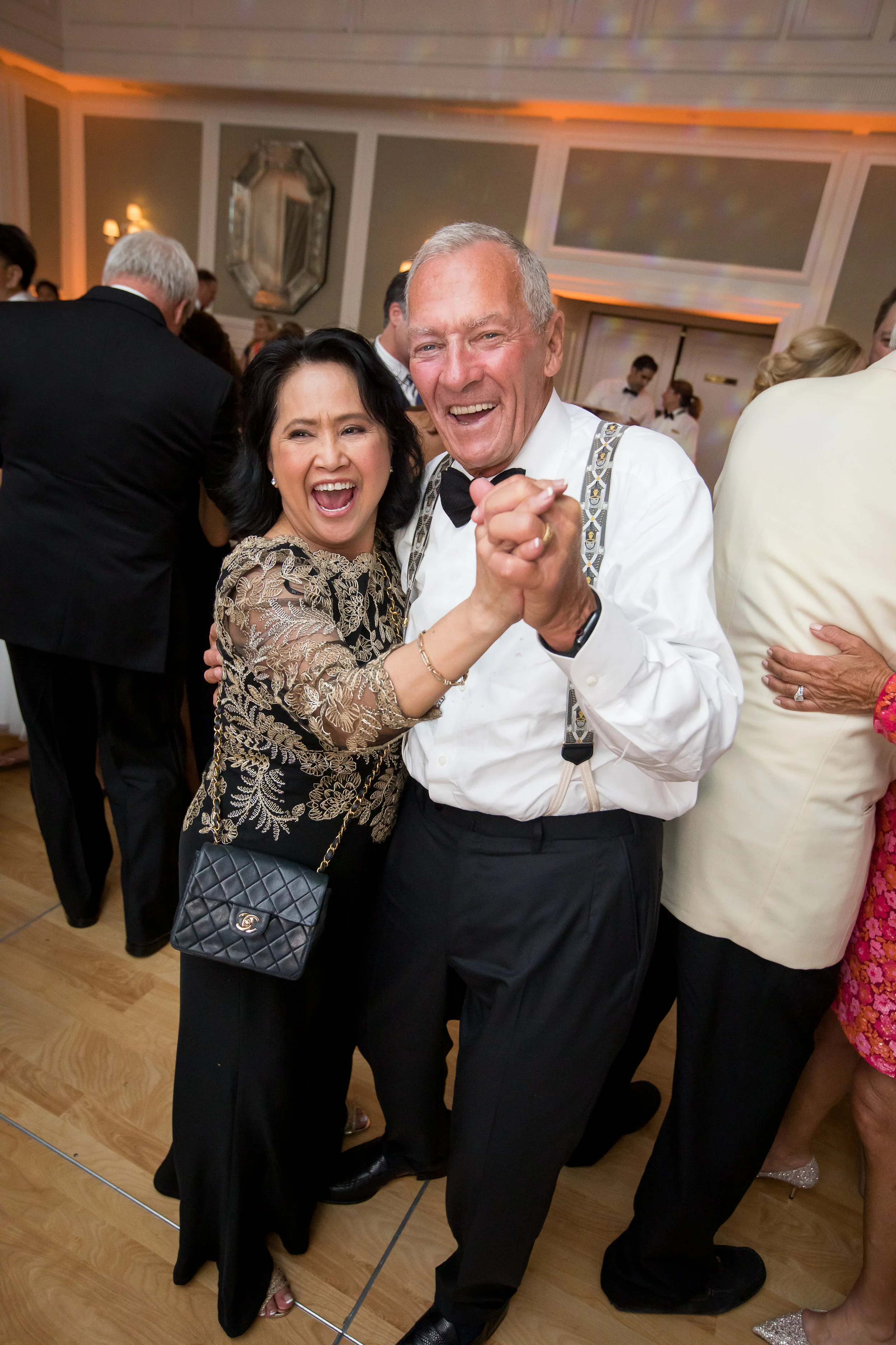A happy couple dancing and celebrating at a formal event or party with other guests in the background.