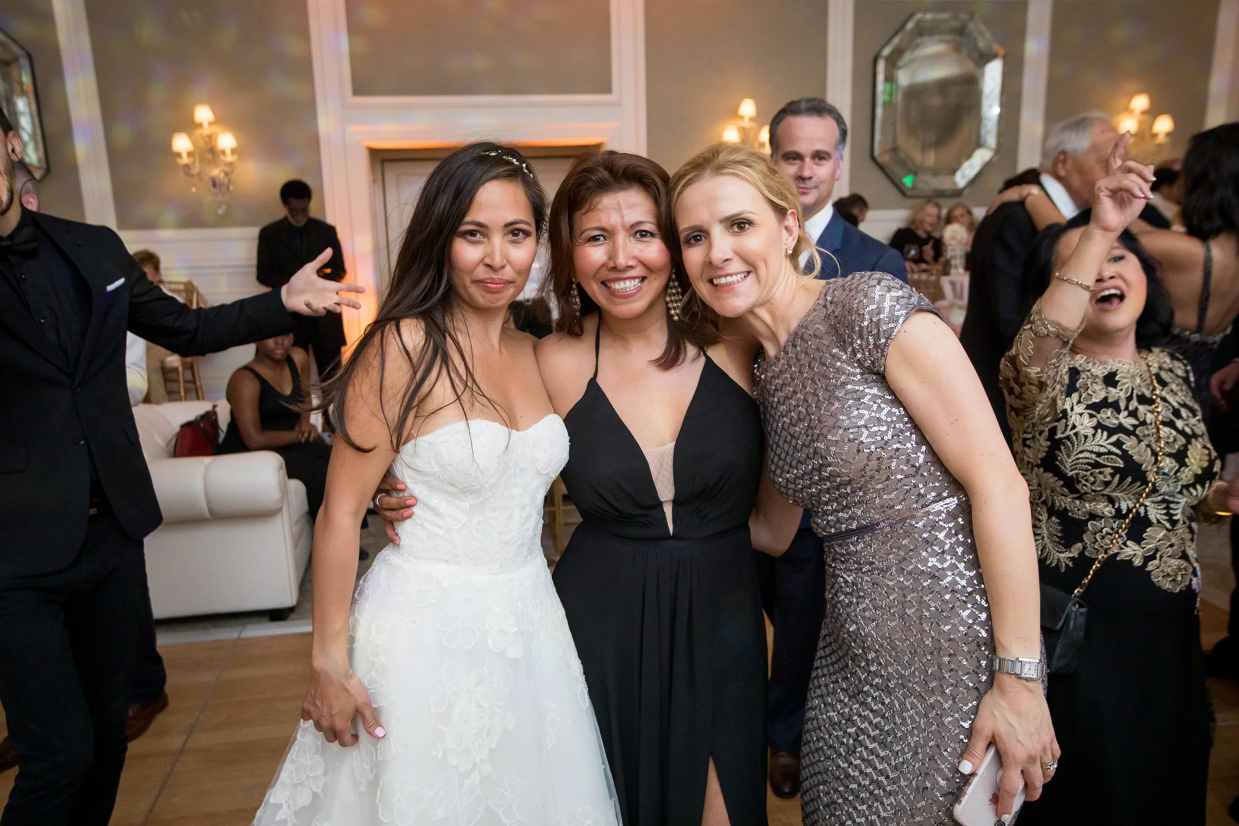 Three women in formal attire smiling and posing together at a celebration or wedding reception.