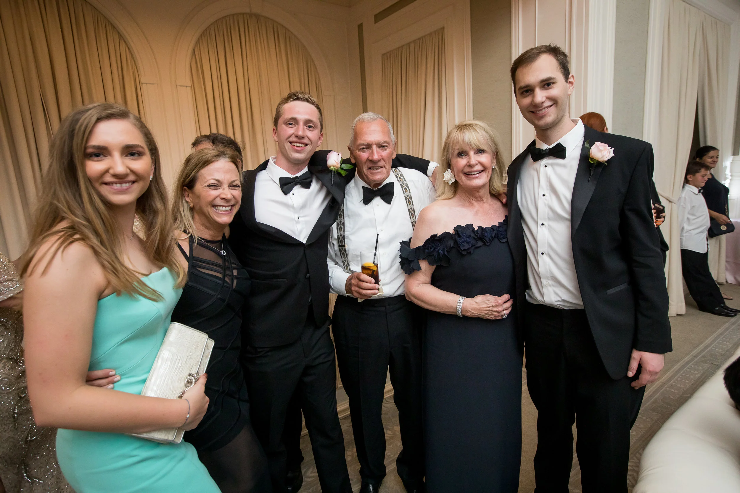 Group of seven people at a formal event, smiling, dressed in tuxedos and an evening gown, standing in a room with cream-colored curtains.