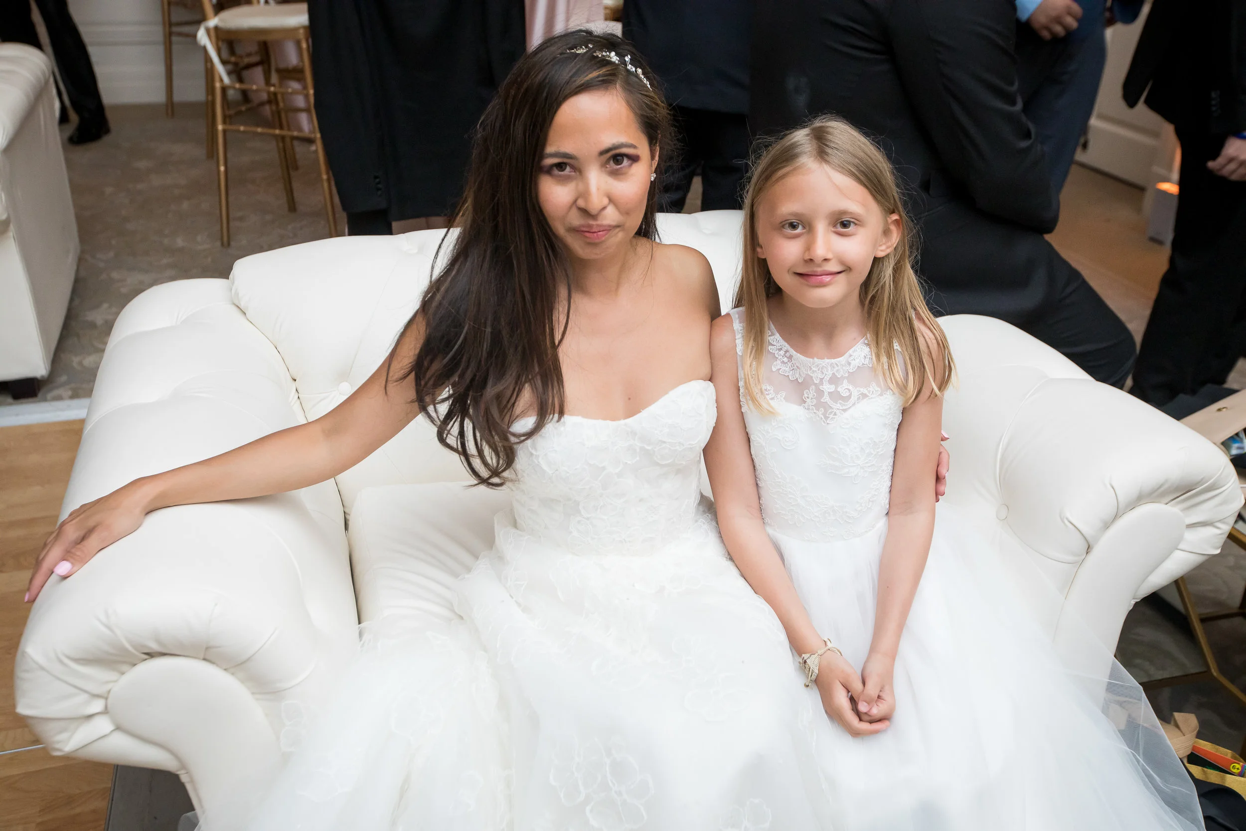 A woman and a young girl sitting on a white sofa at a wedding reception, both dressed in white dresses, with other guests in the background.
