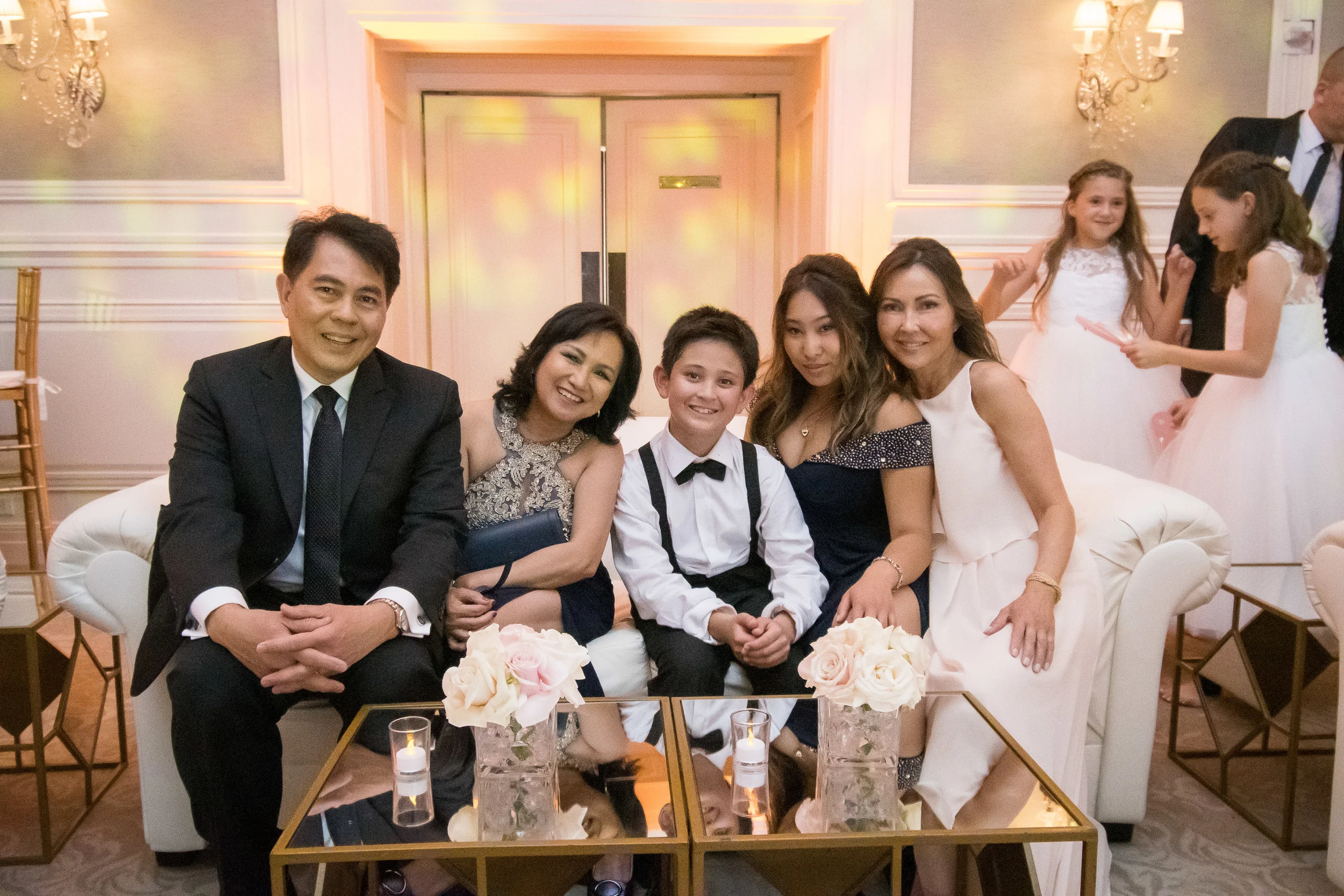 Group of five people sitting on a white sofa at a formal event, with two girls in dresses and a boy in a tuxedo in the background.