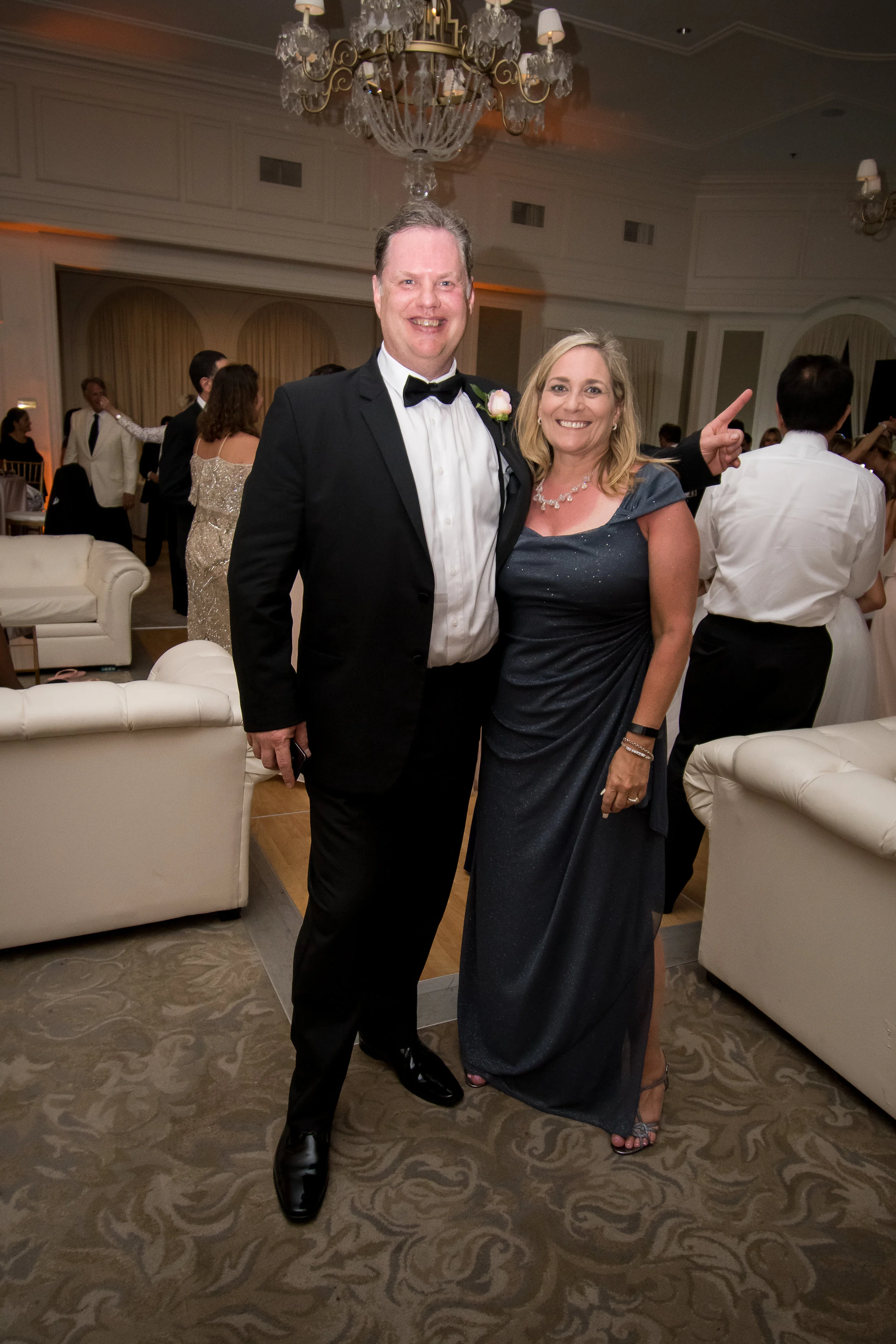 A man in a tuxedo and a woman in a formal dress posing together at a wedding reception, with other guests and a chandelier in the background.