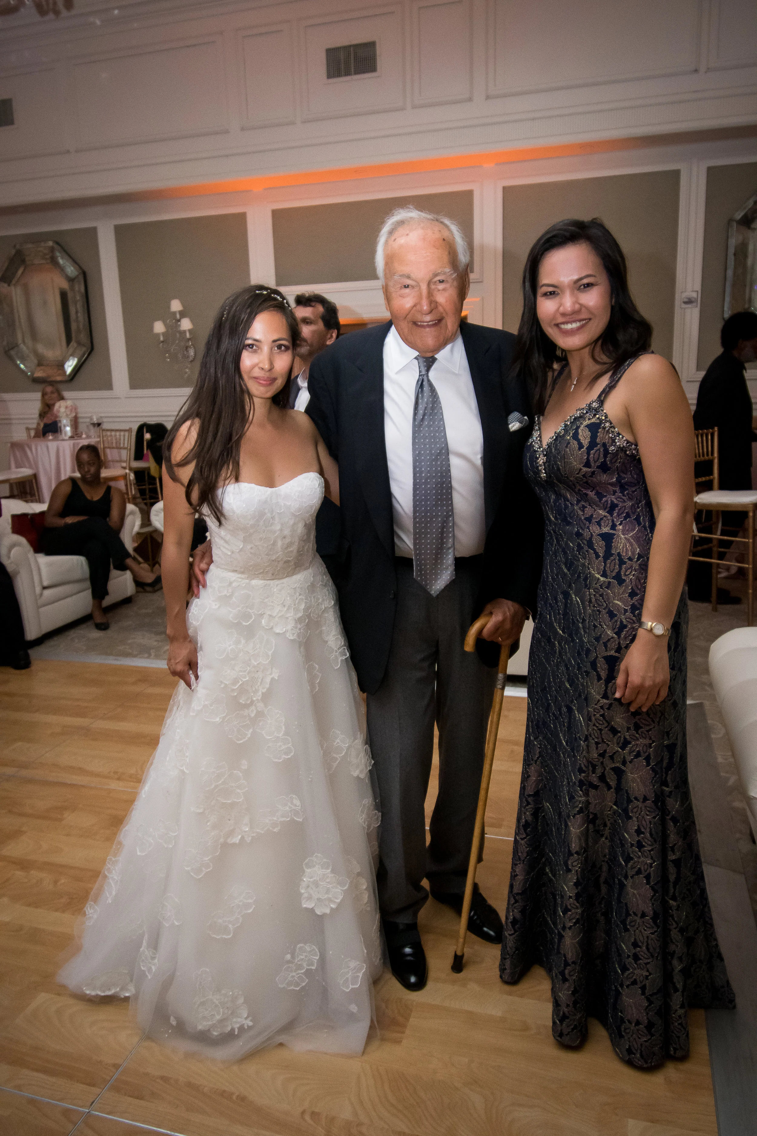 Three people standing together at a celebration: a young woman in a white wedding dress, an elderly man with a cane wearing a dark suit, and a young woman in a dark patterned gown, smiling indoors.