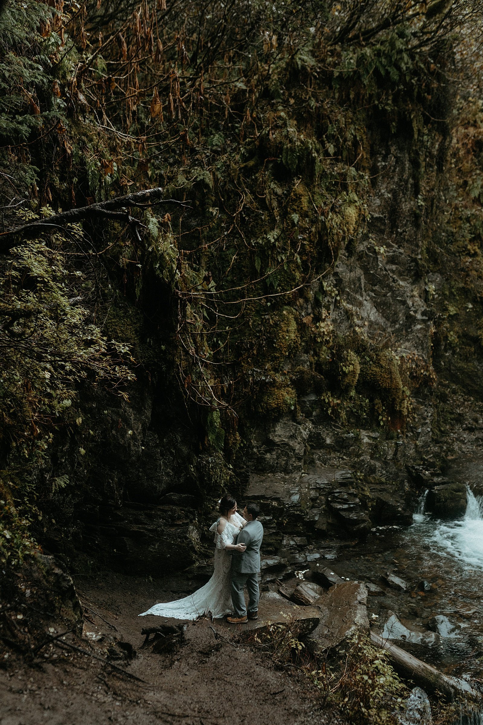  Newlyweds under a lush green waterfall in the Alaska woods 