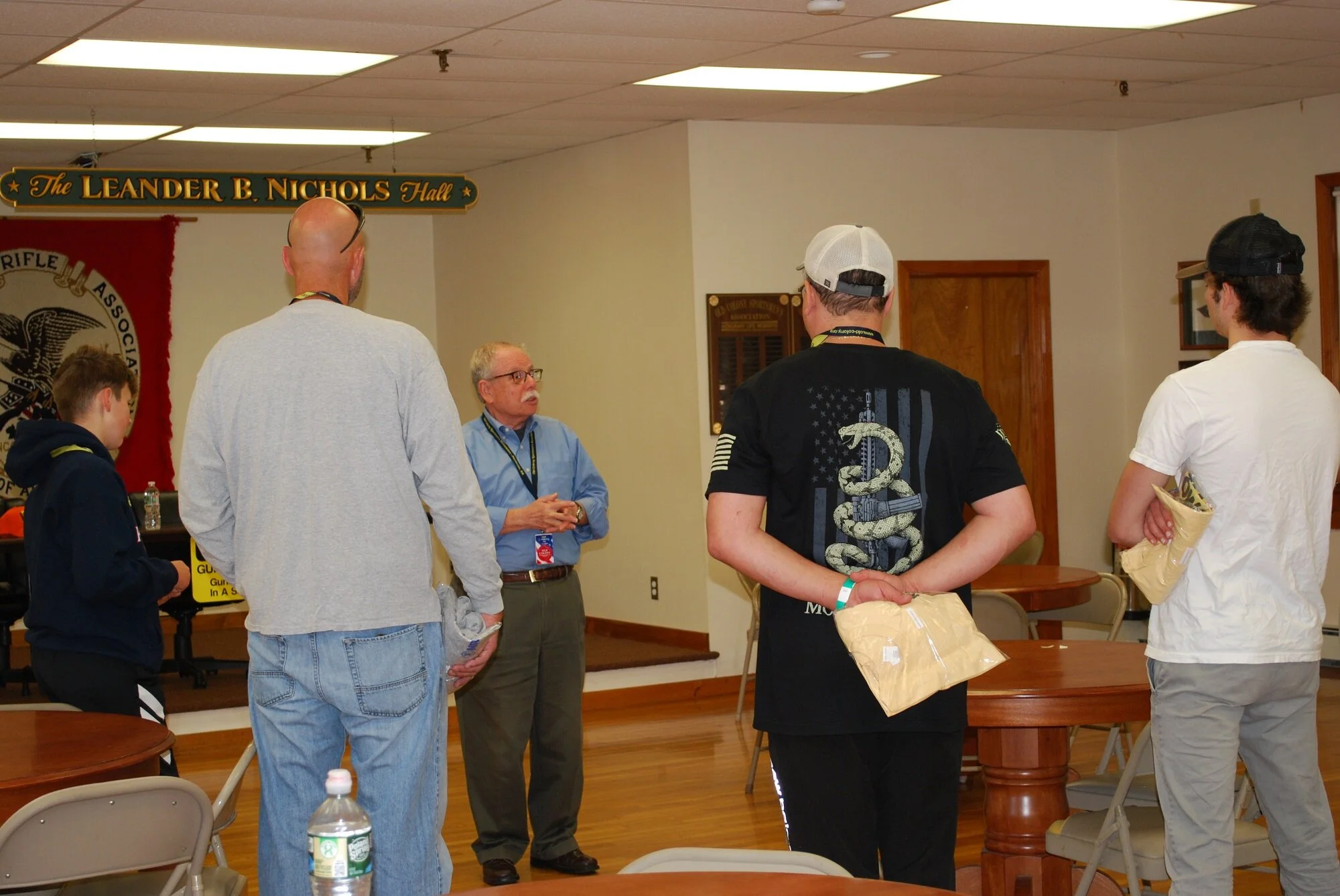 A group of people standing and talking in a room with wood floors and a sign that says 'The Leander B. Nichols Hall', with a banner displaying an eagle insignia on the wall.