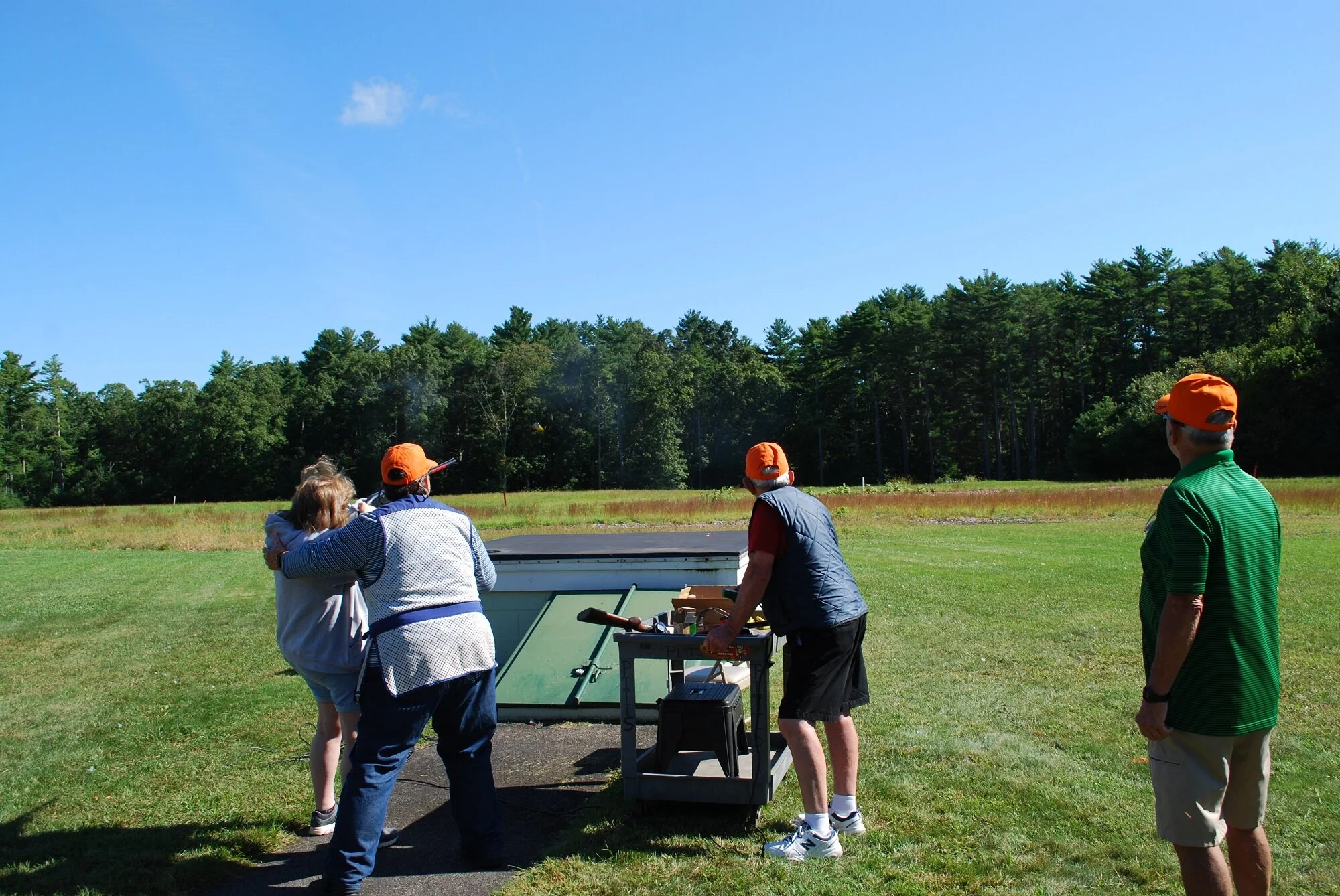 Group of five people, three wearing orange caps, gathered outdoors on a grassy field with trees in the background, participating in a clay shooting activity, with a person aiming a shotgun.