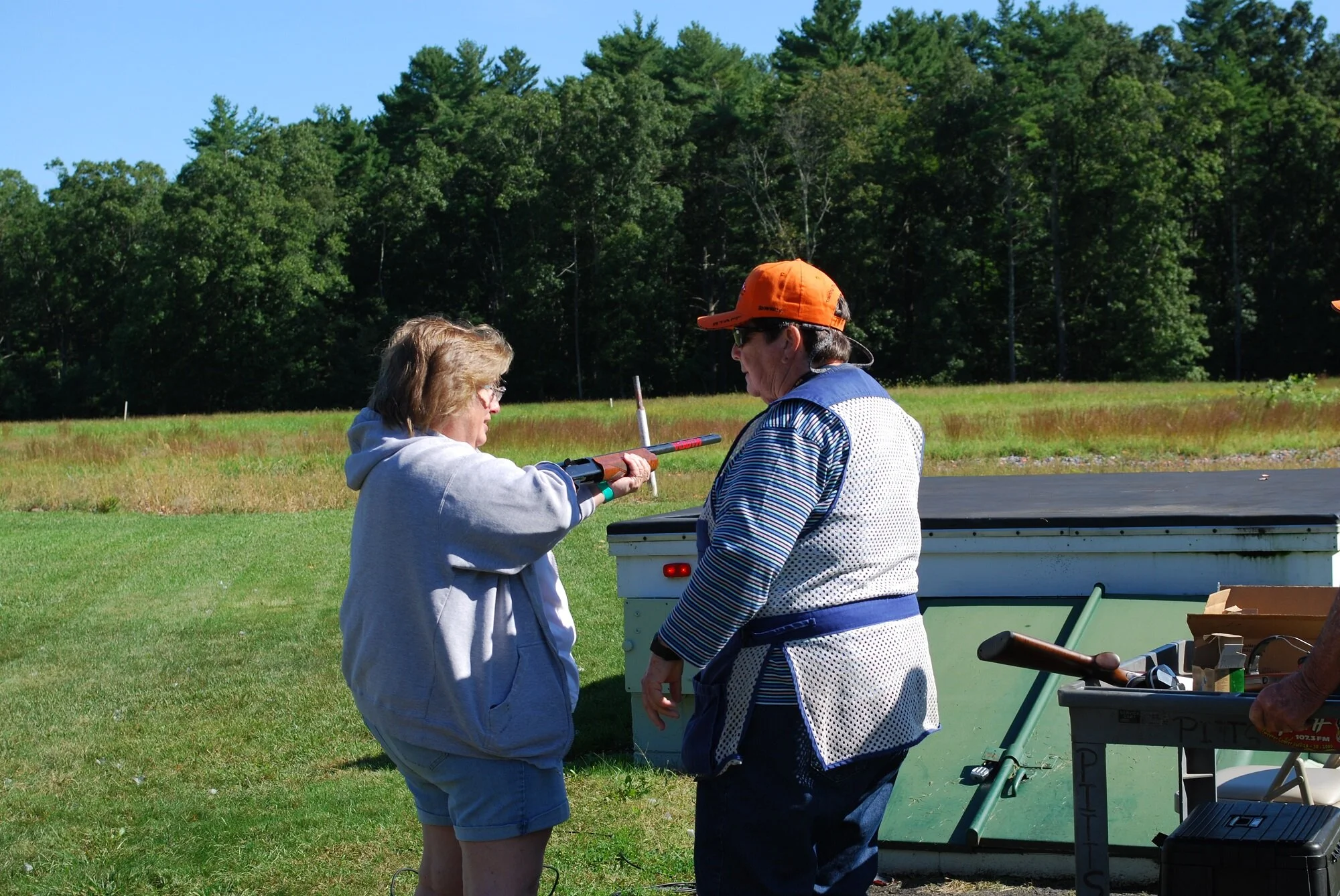 Two women outdoors on a grassy field, one handing a shotgun to the other, with trees in the background.