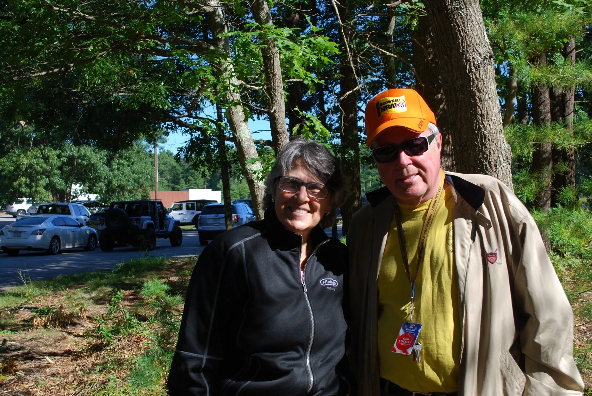 Two people standing outdoors in front of trees, smiling at the camera. The person on the left is a woman with short dark hair, wearing glasses and a black jacket. The person on the right is a man with light hair, wearing sunglasses, an orange cap wit