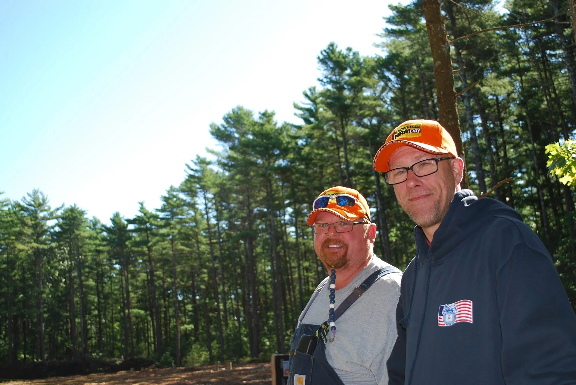 Two men in orange hats and glasses standing outdoors in a forest with tall trees and green foliage, smiling at the camera.