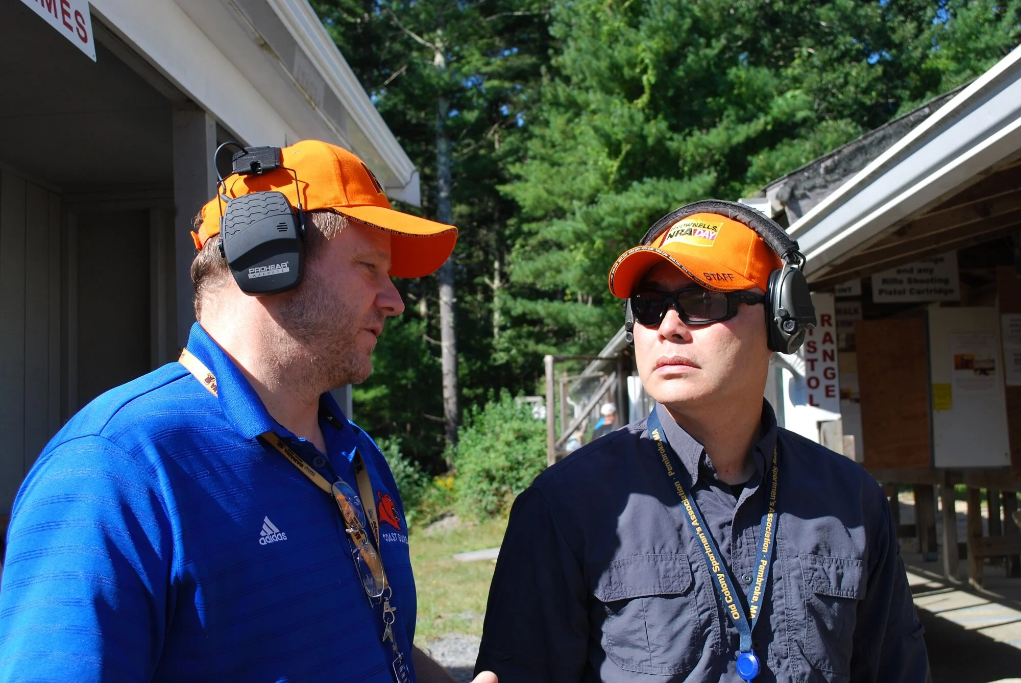Two men wearing orange caps, sunglasses, and headsets standing outside near a building, engaged in conversation. One is in a blue shirt with a logo, the other in a dark gray shirt. Green trees in the background.