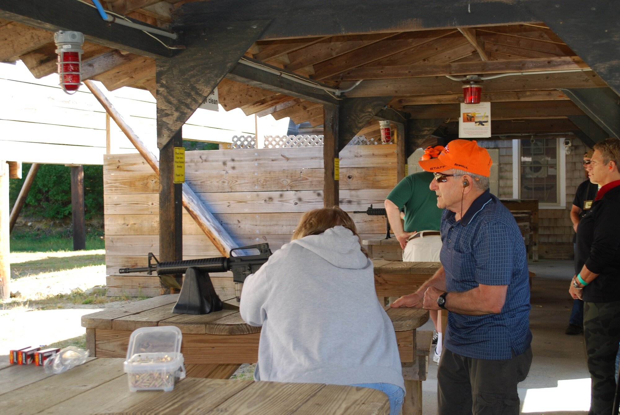 People at an outdoor shooting range with a rifle on a wooden table, engaged in a firearms training or practice session.